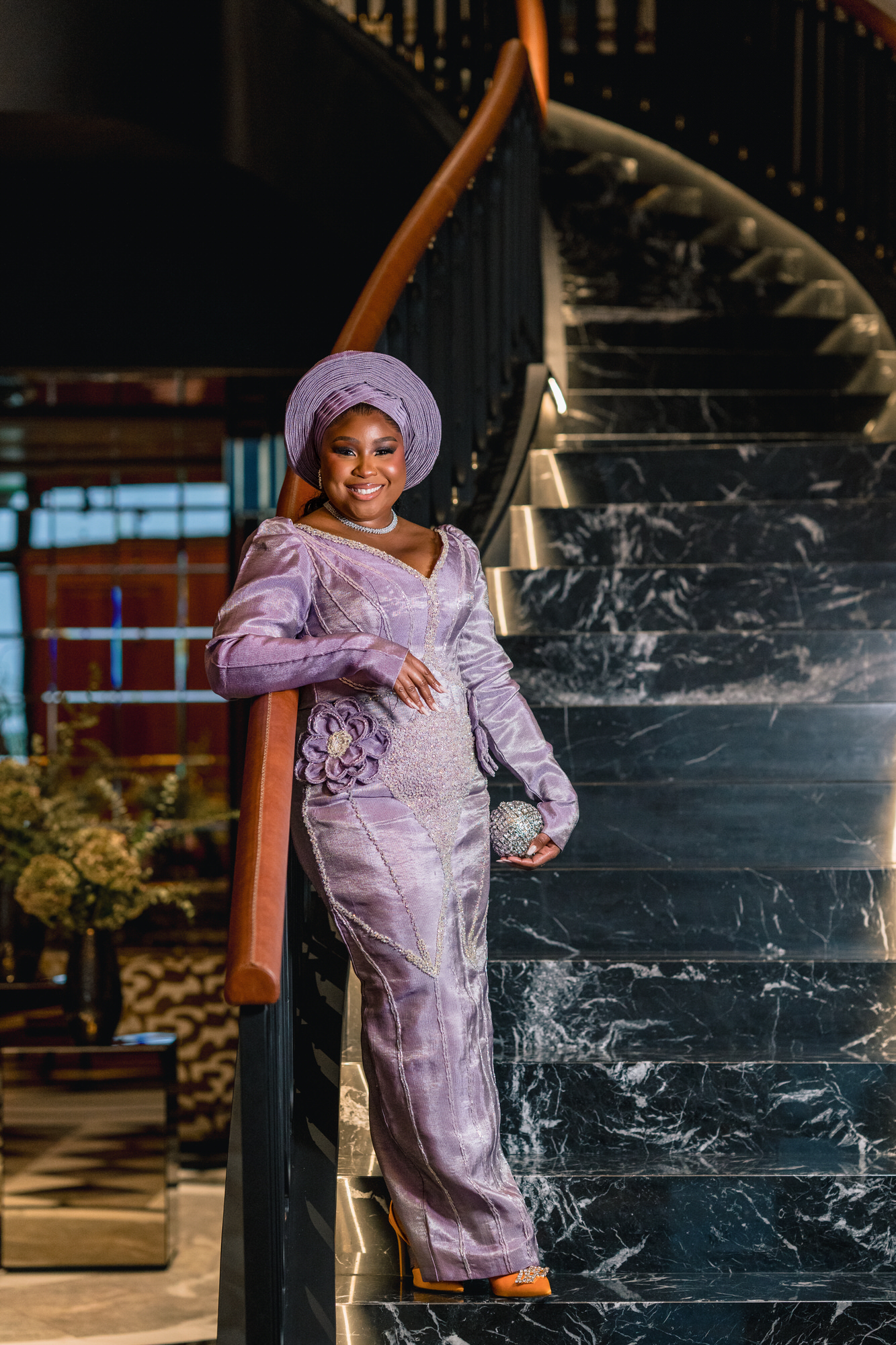 Smiling woman in a lavender traditional dress and matching headwrap poses on a black marble staircase indoors, holding a beaded clutch.