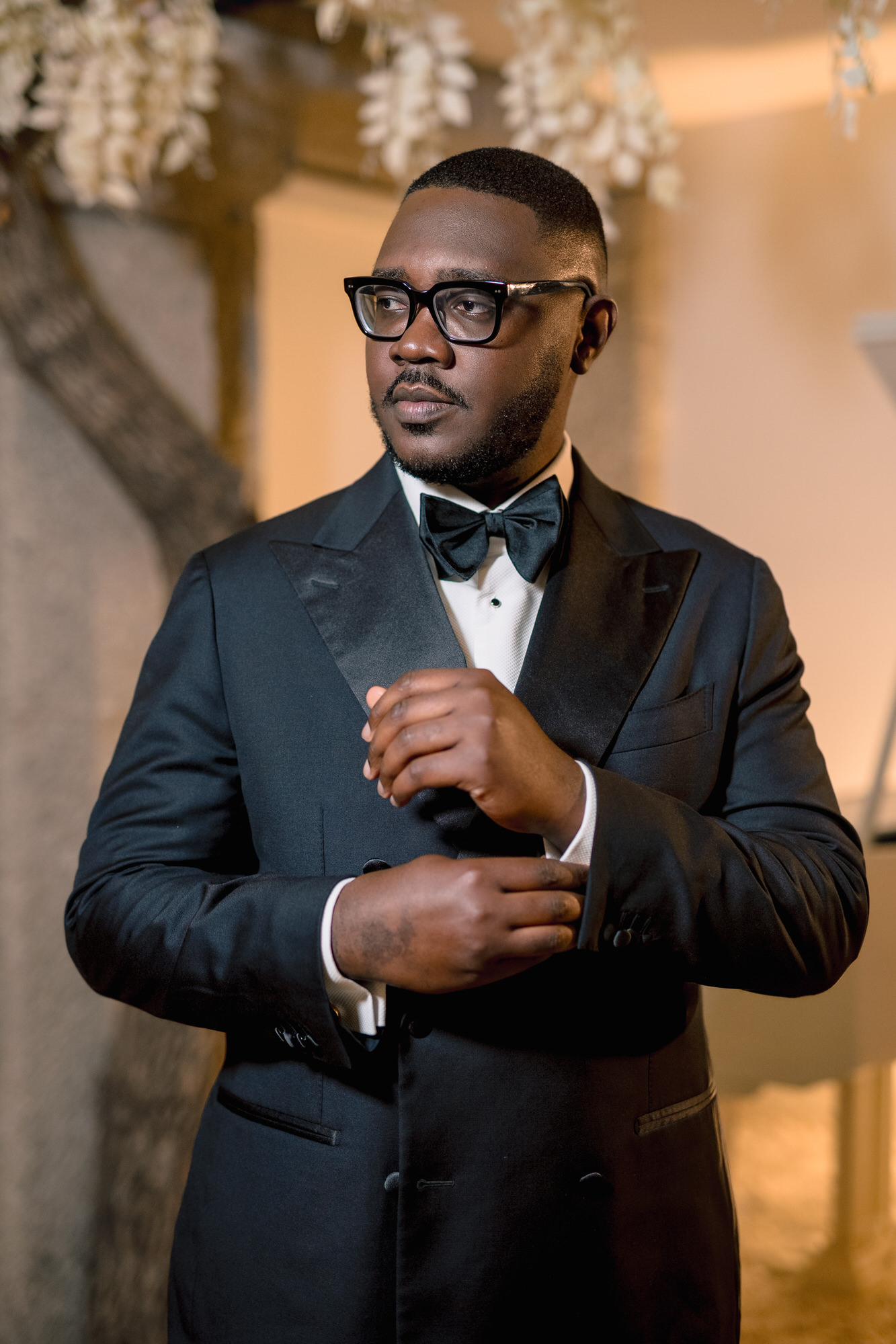 Man in a black tuxedo and bow tie adjusts his cuff while wearing glasses in a warmly lit indoor setting.
