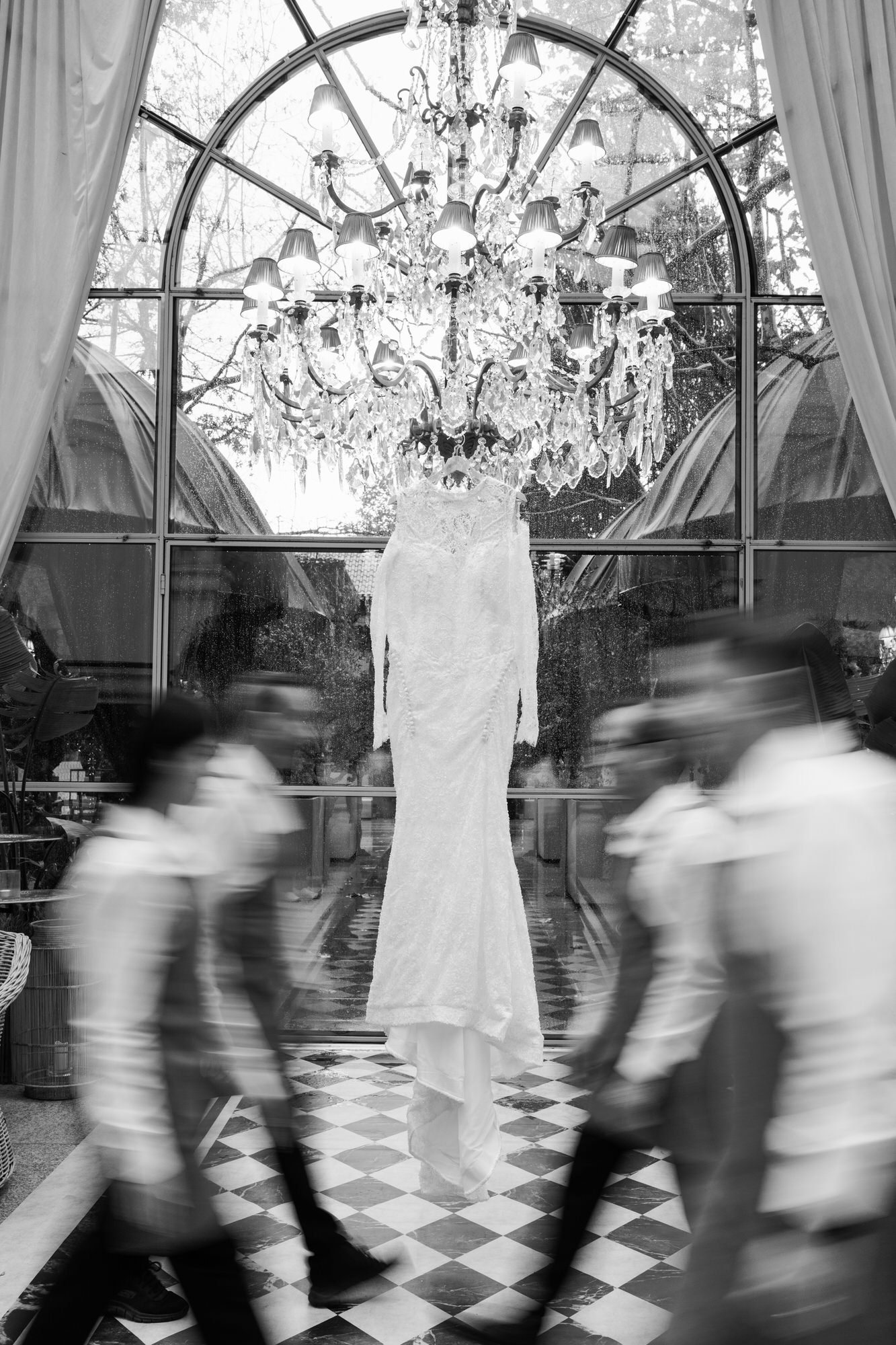 Wedding dress on a mannequin inside a glass-walled venue with a grand chandelier, while blurred guests pass by in the foreground.