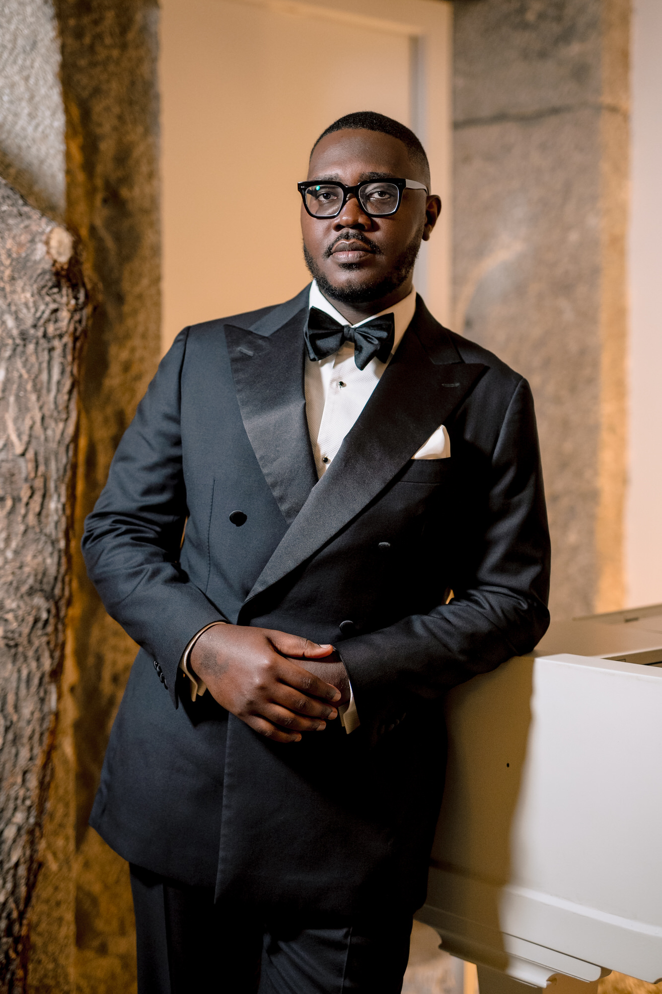 Formal portrait of a man in a black tuxedo, bow tie, and glasses, standing beside a piano near a stone wall.