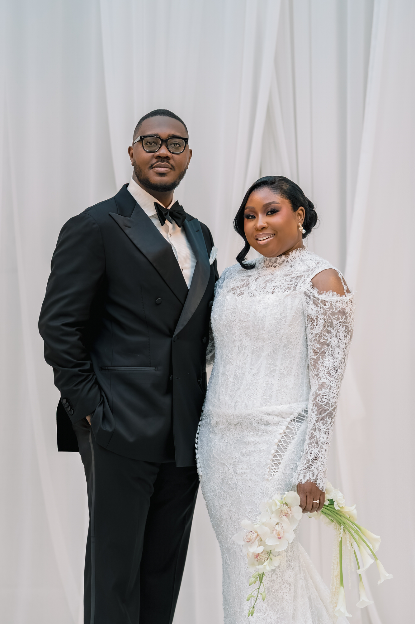 Bride in a lace wedding gown stands beside a groom in a black tuxedo against a white backdrop, posing for a wedding photo.