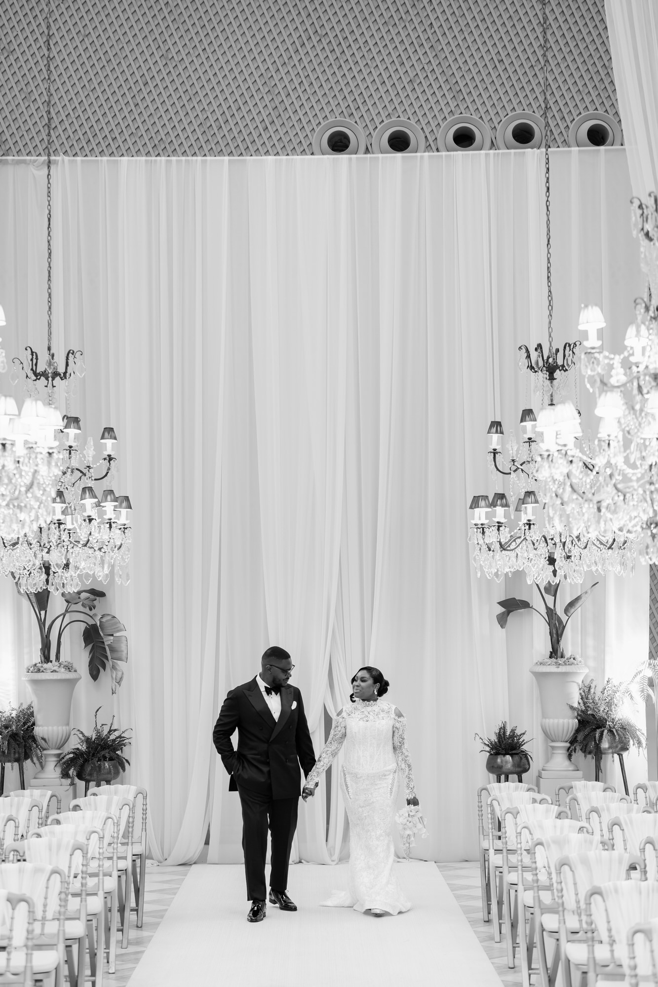 Bride in a white lace gown and groom in a black tux walk hand in hand down a white aisle under draped backdrop and chandeliers at a wedding venue.