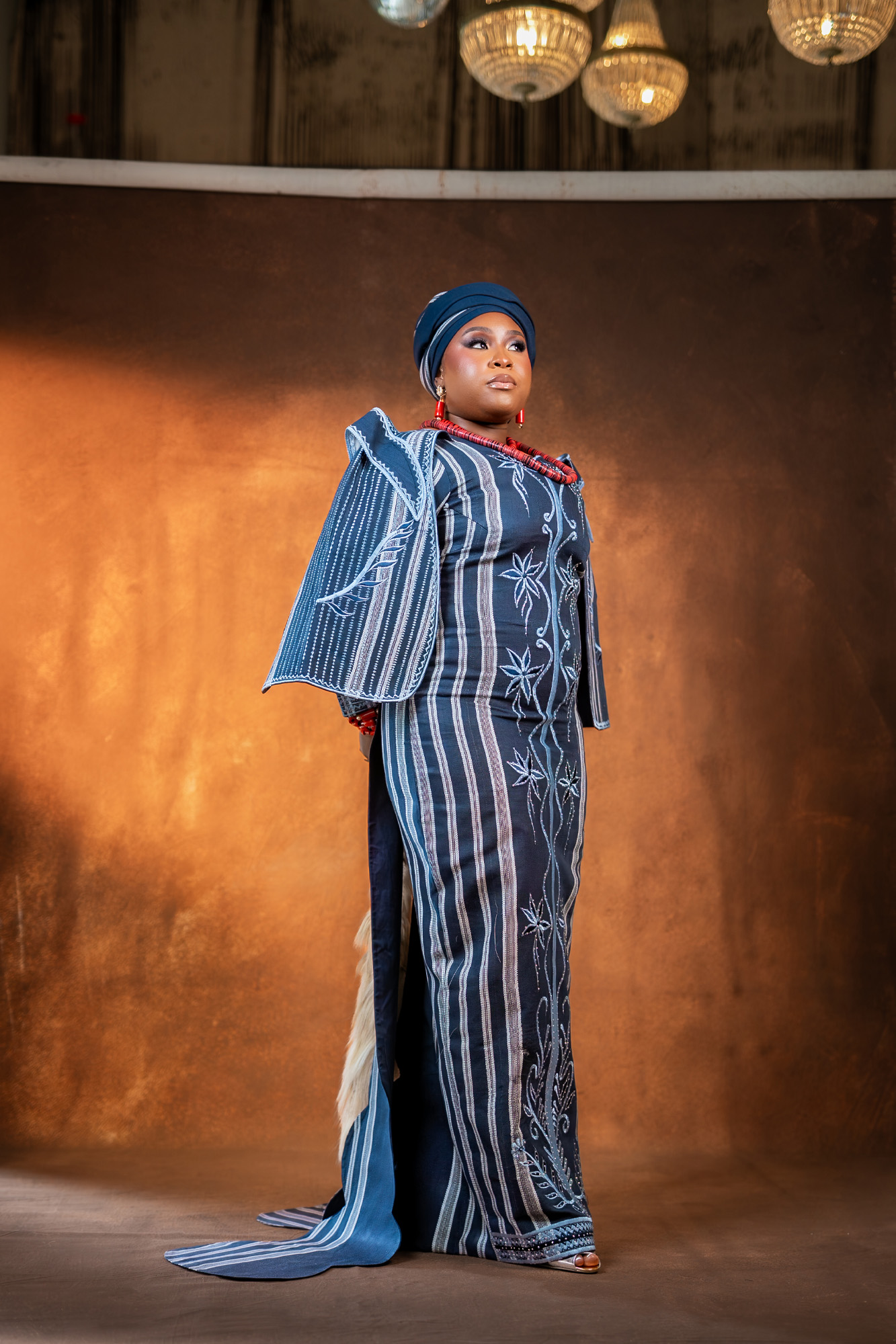 Full-length portrait of a woman in a blue patterned traditional dress with matching headwrap, standing against a warm brown backdrop.