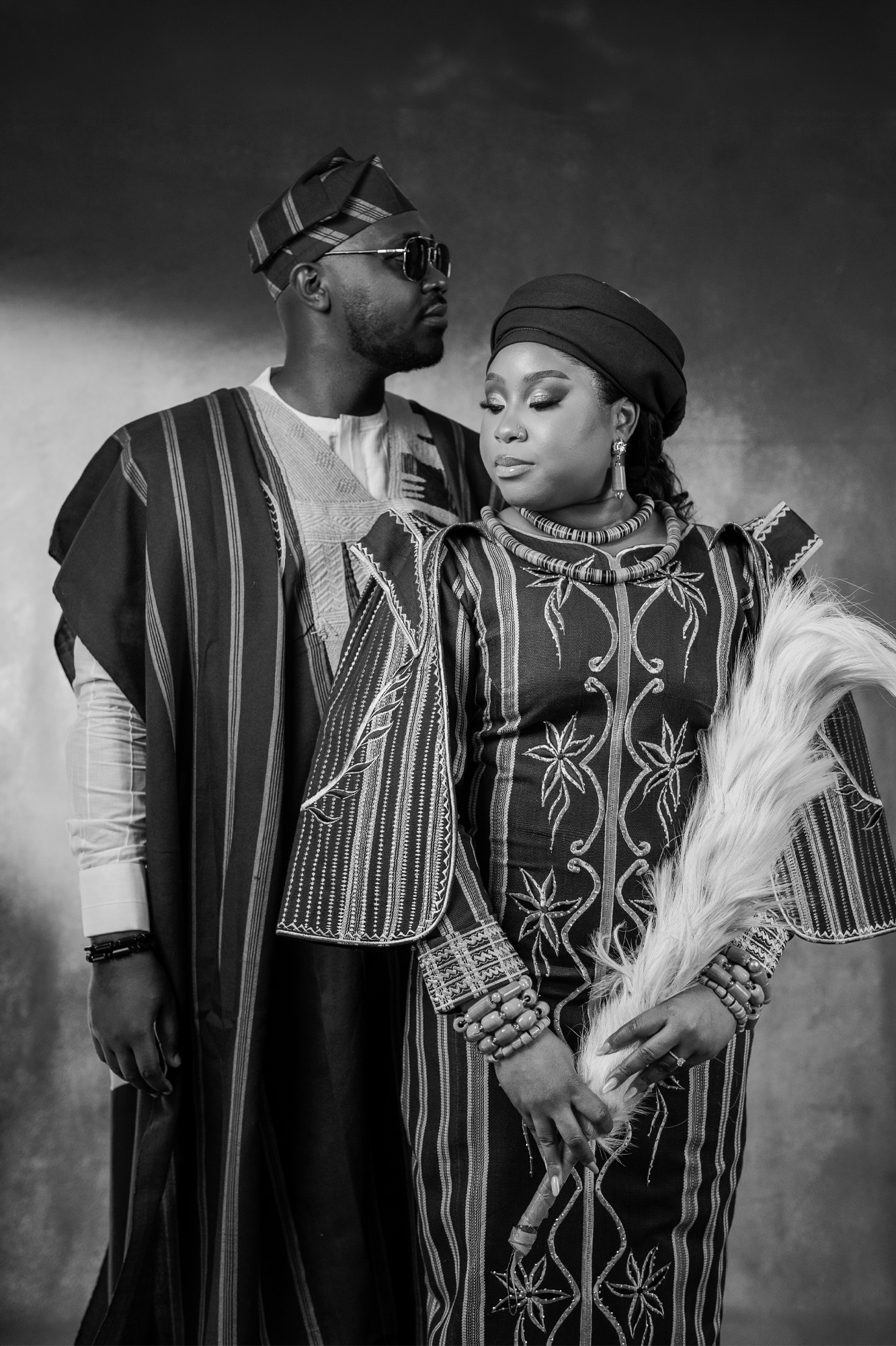 Couple in traditional African attire posing together; man wears a striped agbada and cap, woman wears an embroidered dress with a head wrap, holding a feathered accessory.