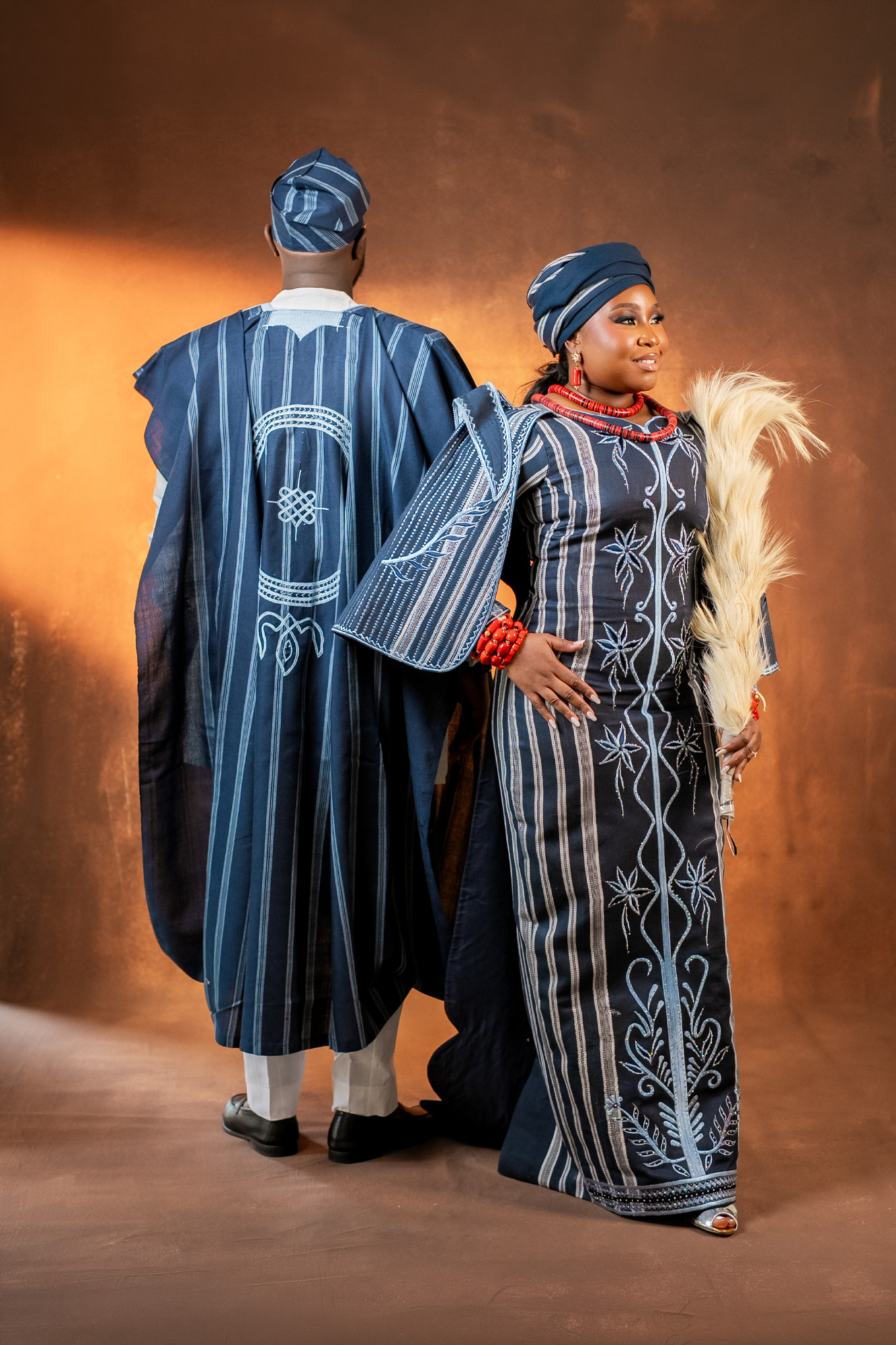 Man in a blue striped traditional robe stands with his back to the camera, while a smiling woman in a matching blue dress and head wrap faces forward, holding a feathered stole and wearing red jewelry against a bronze backdrop.