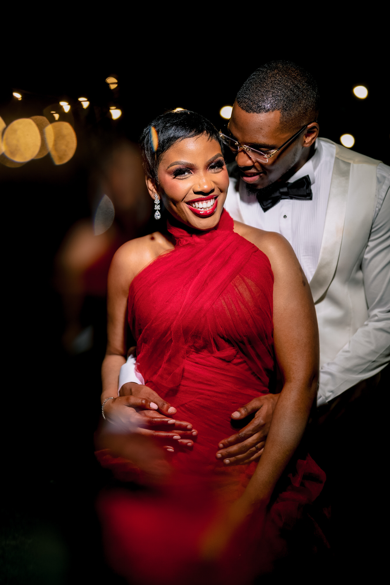 Couple dancing at night: woman in a red dress smiling, man in a white tuxedo with a bow tie standing close behind her; warm bokeh lights in the background.