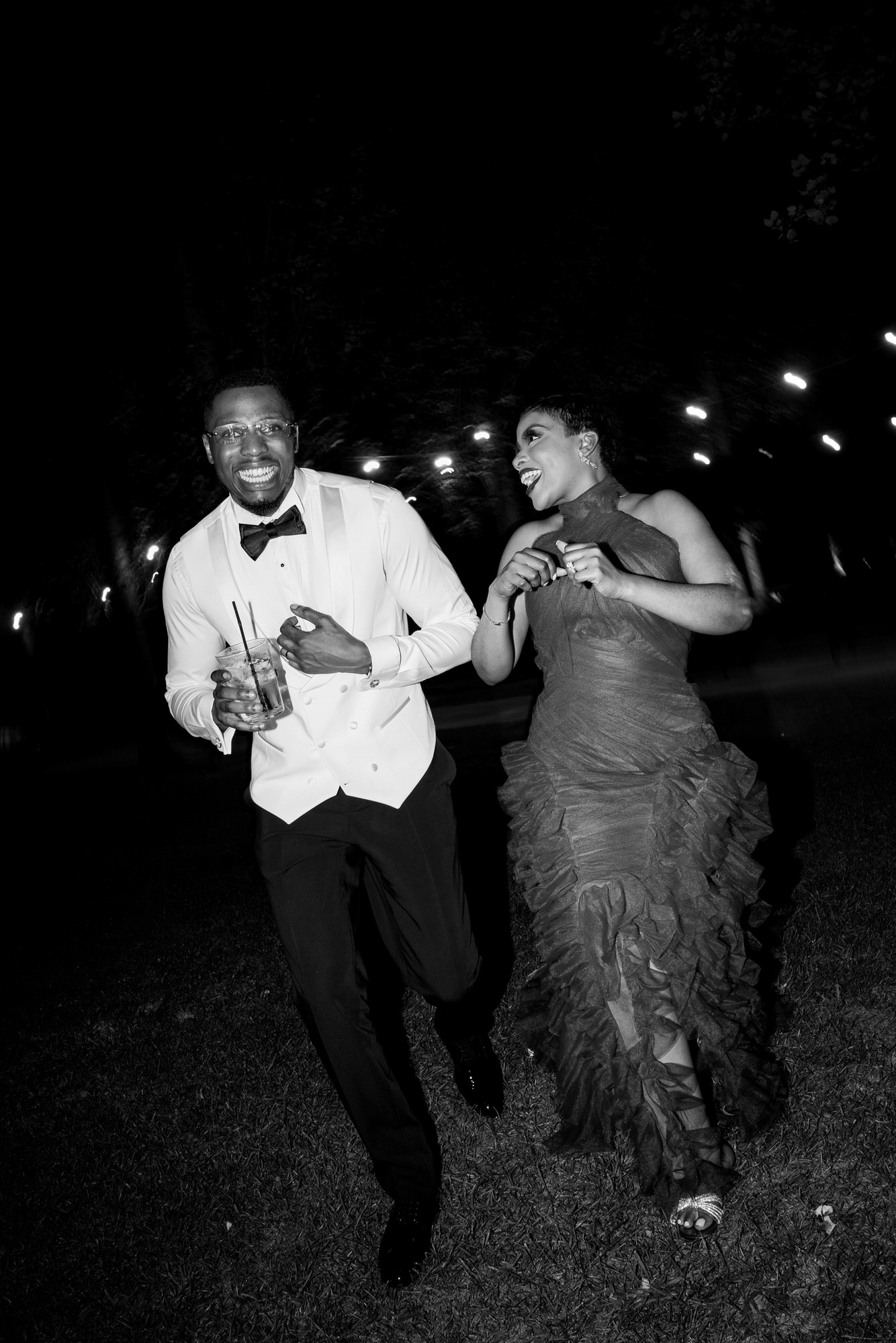 Bride in a lace wedding gown and veil, holding a bouquet of white flowers, next to a groom in a white tuxedo outdoors.