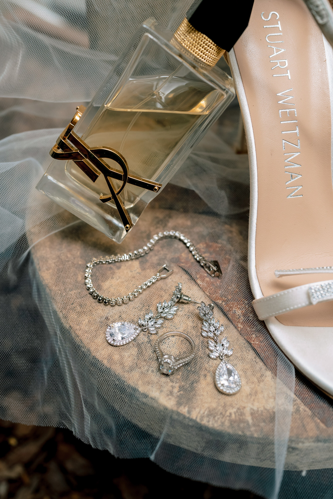 Jewelry pieces—diamond necklace, earrings, and bracelet laid on sheer fabric beside a pale pink Stuart Weitzman shoe heel