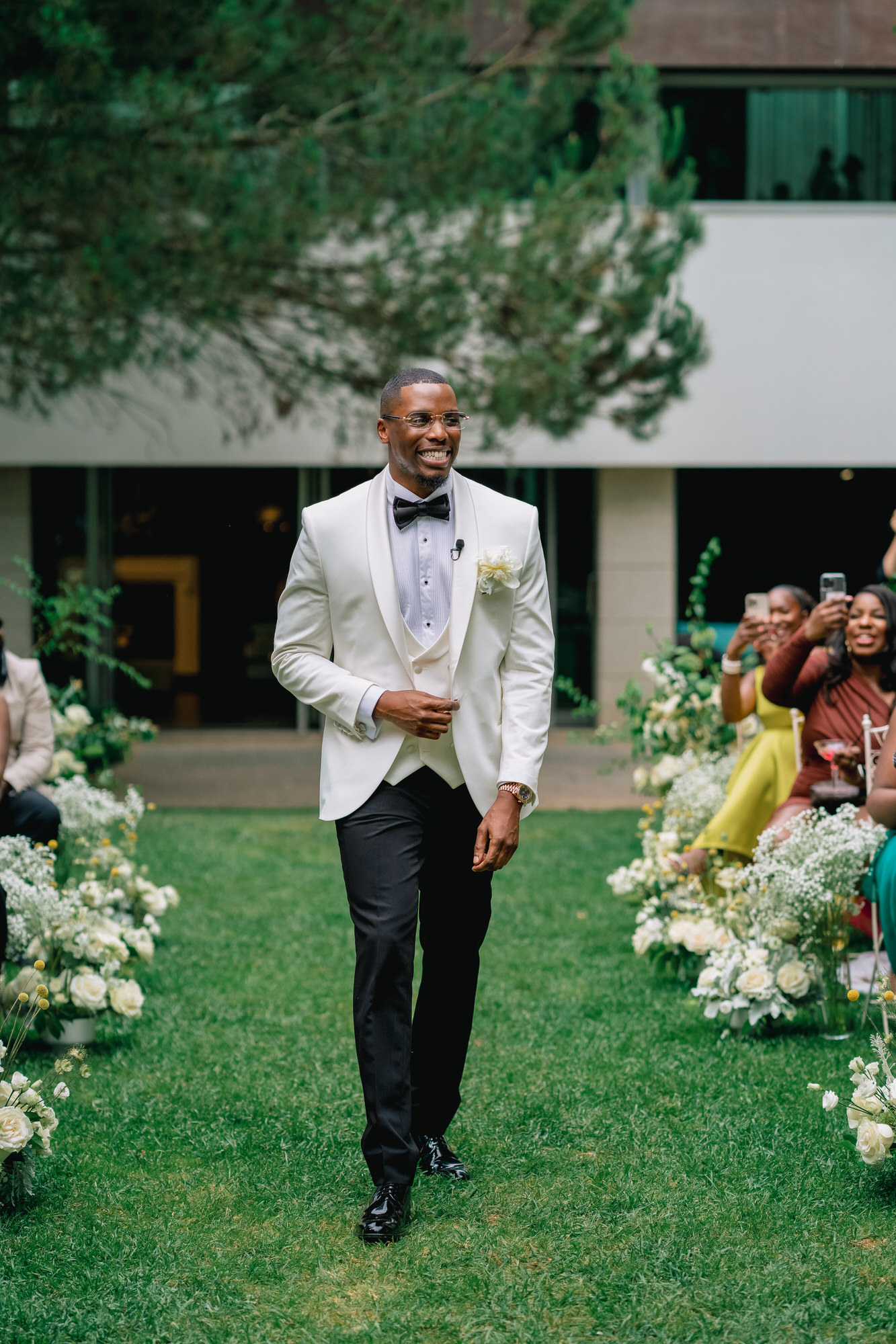 Groom in a white dinner jacket and black bow tie walking down a grassy outdoor wedding aisle with guests taking photos in the background.