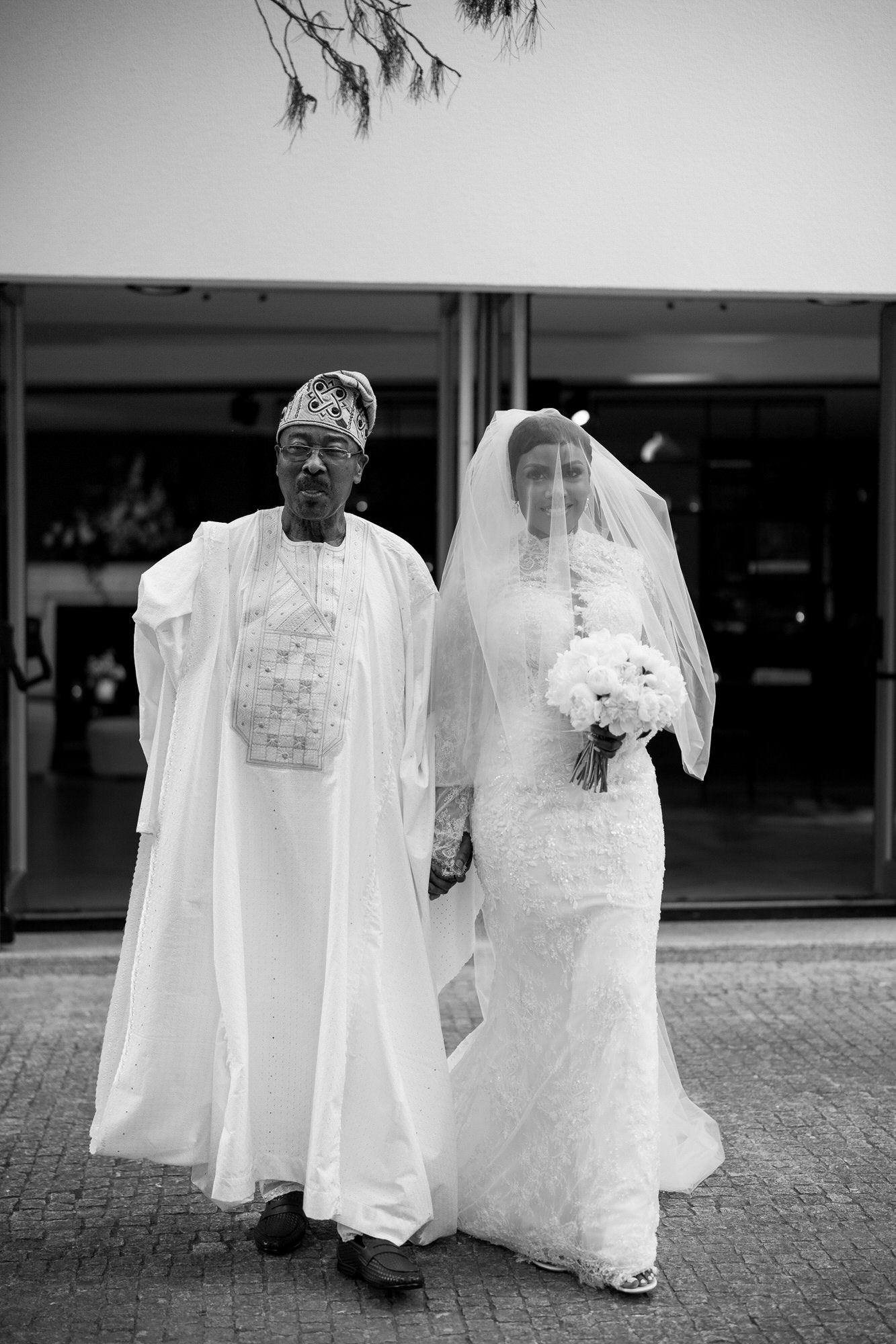 Bride in a lace wedding gown and veil holds a bouquet while walking with a man in traditional white agbada outside a building (black and white).