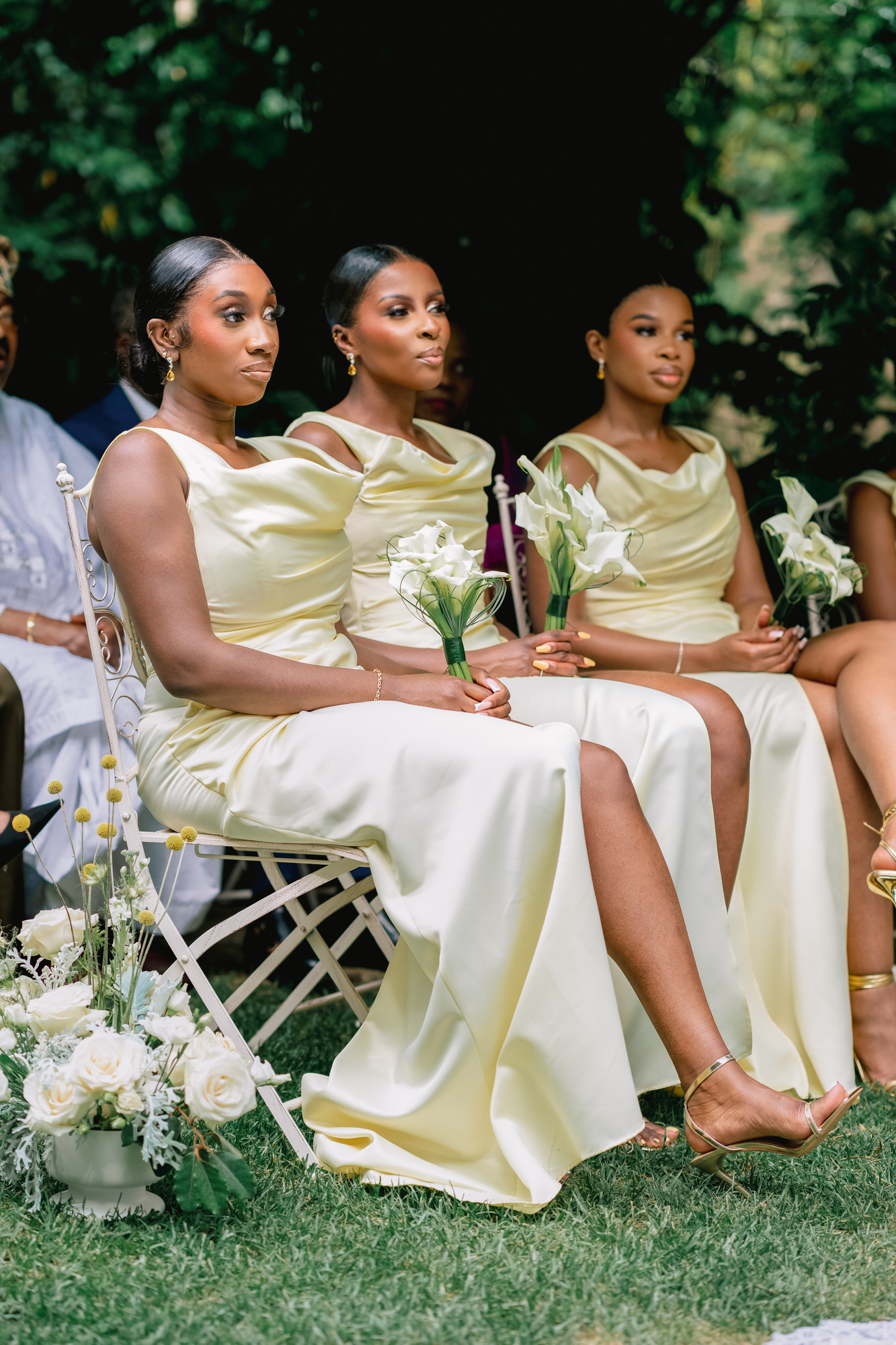 Three women in pale yellow bridesmaid dresses sit on white chairs outdoors, holding white bouquets with a green garden backdrop.