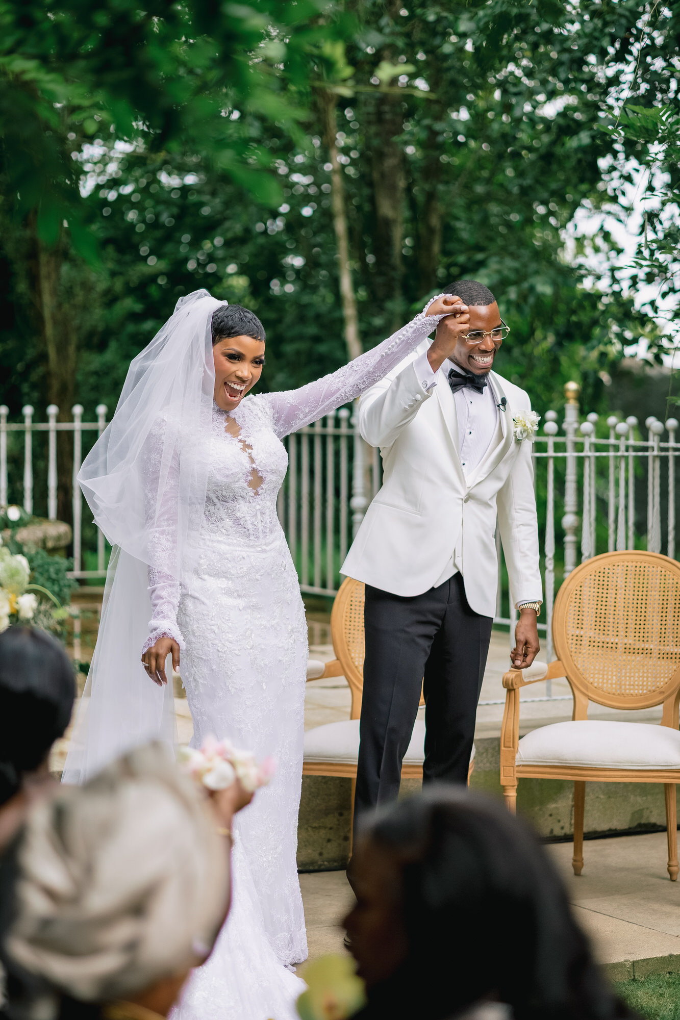 Bride and groom smiling and dancing hand-in-hand outdoors at a wedding, in a garden setting with white chairs nearby