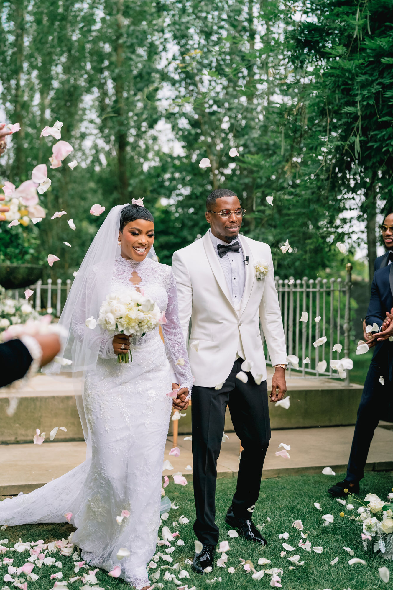 Bride in a lace wedding dress and groom in a white suit walk hand in hand as pink petals float around them outdoors.