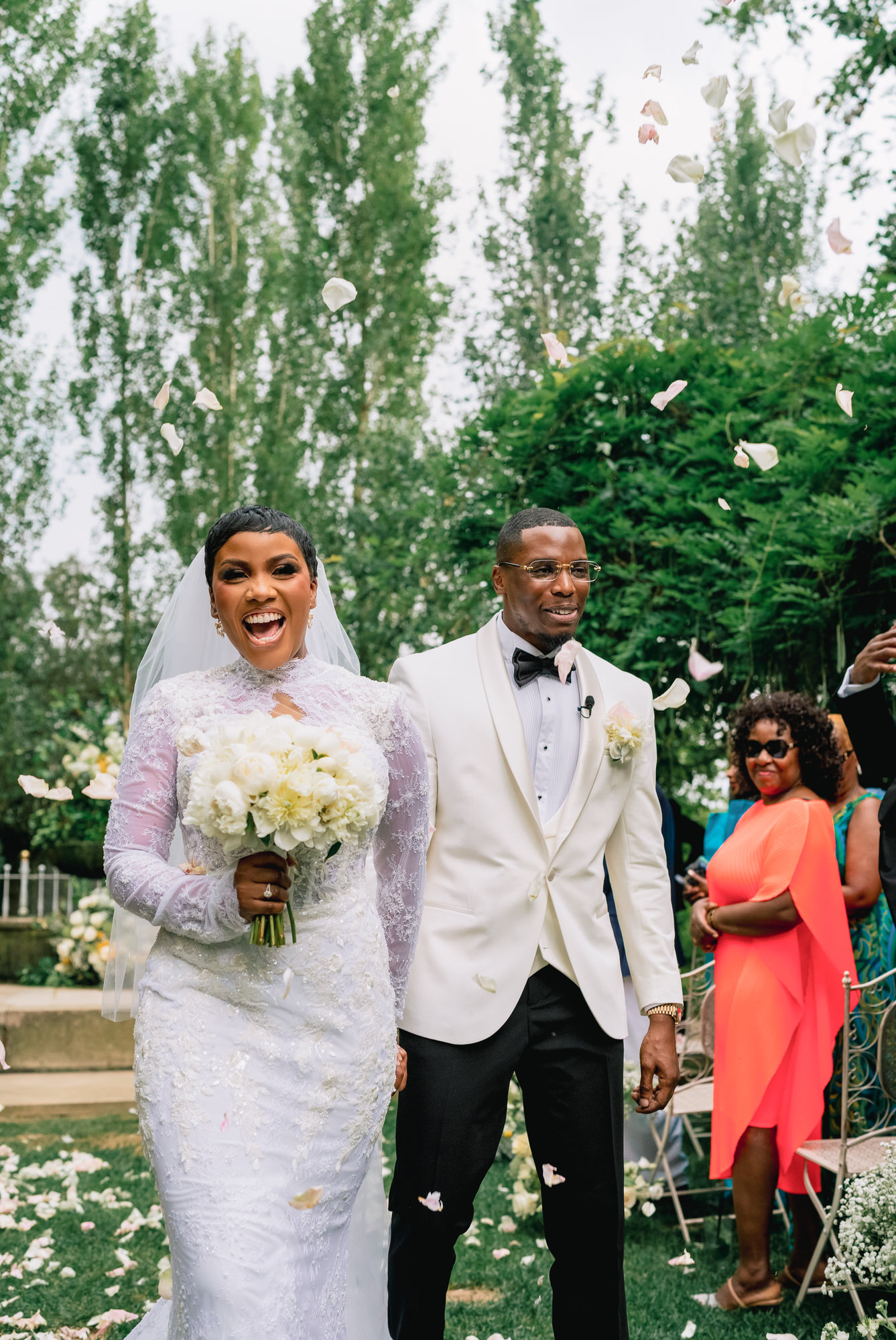 Bride in a white lace wedding gown holding a bouquet, smiling as petals fall around her with a cheering groom beside her outdoors.