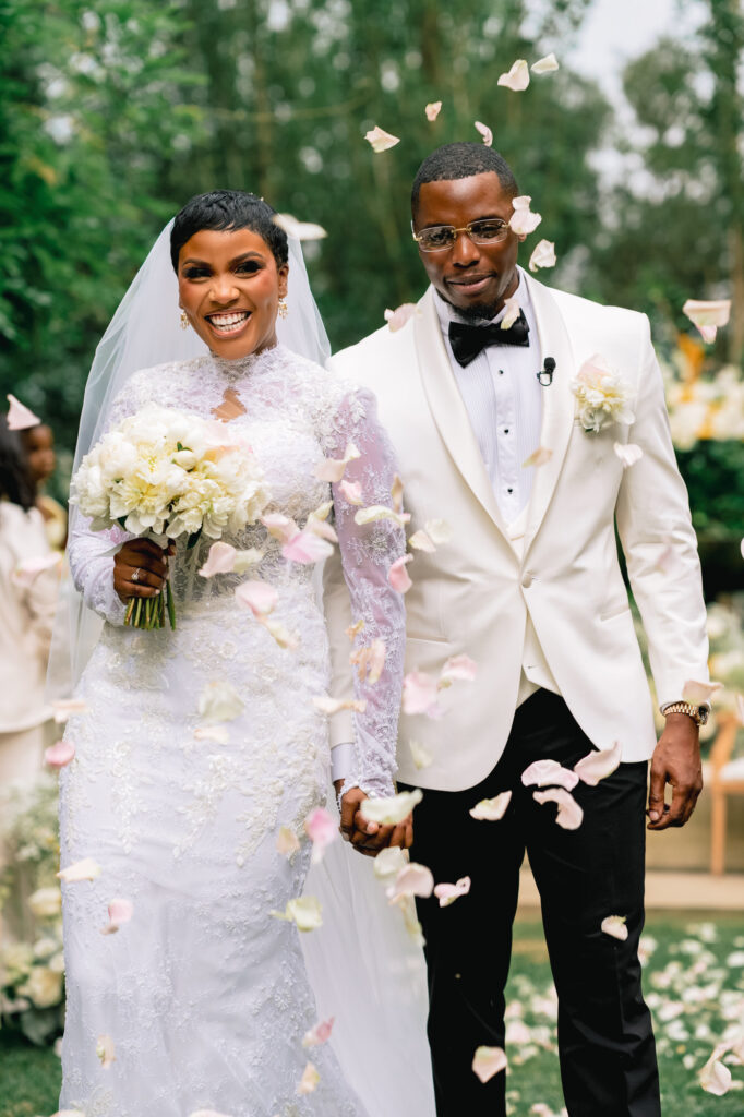 The newlyweds walk hand in hand as pink rose petals drift around them, the bride in a long-sleeve lace gown and veil, the groom in a white dinner jacket and black bow tie.