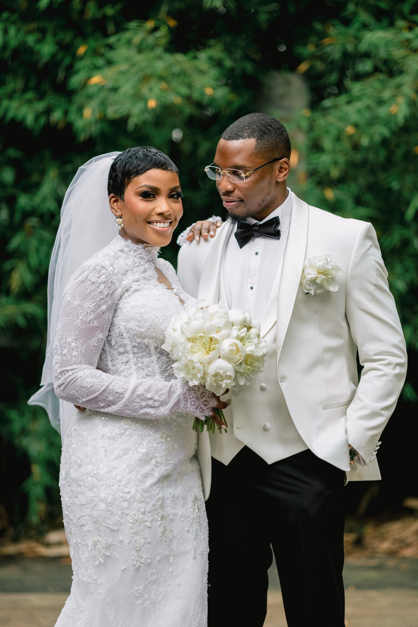 Bride in a lace wedding gown and veil, holding a bouquet of white flowers, next to a groom in a white tuxedo outdoors.