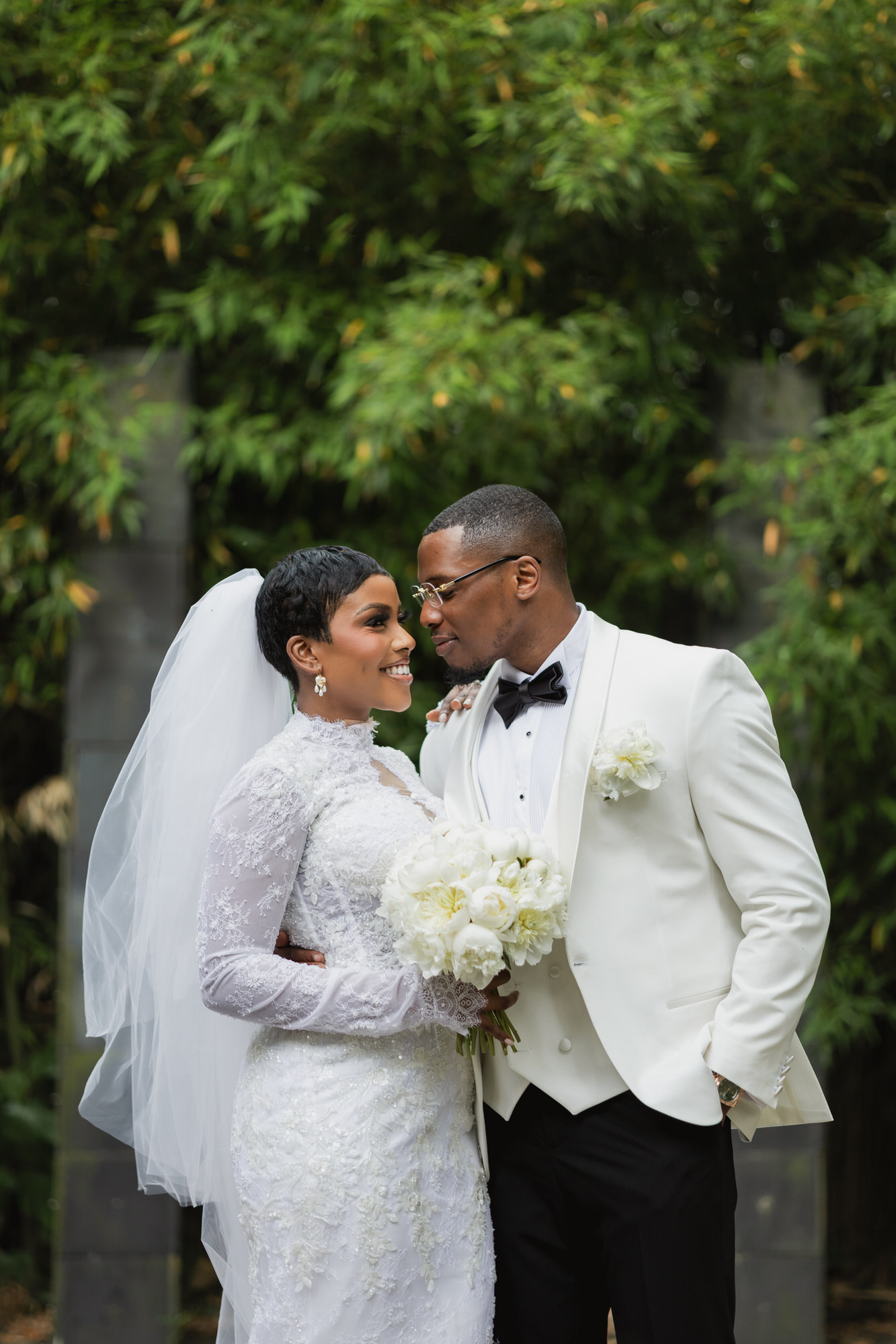Bride in lace wedding gown and veil smiling at groom outdoors, holding a bouquet of white flowers.