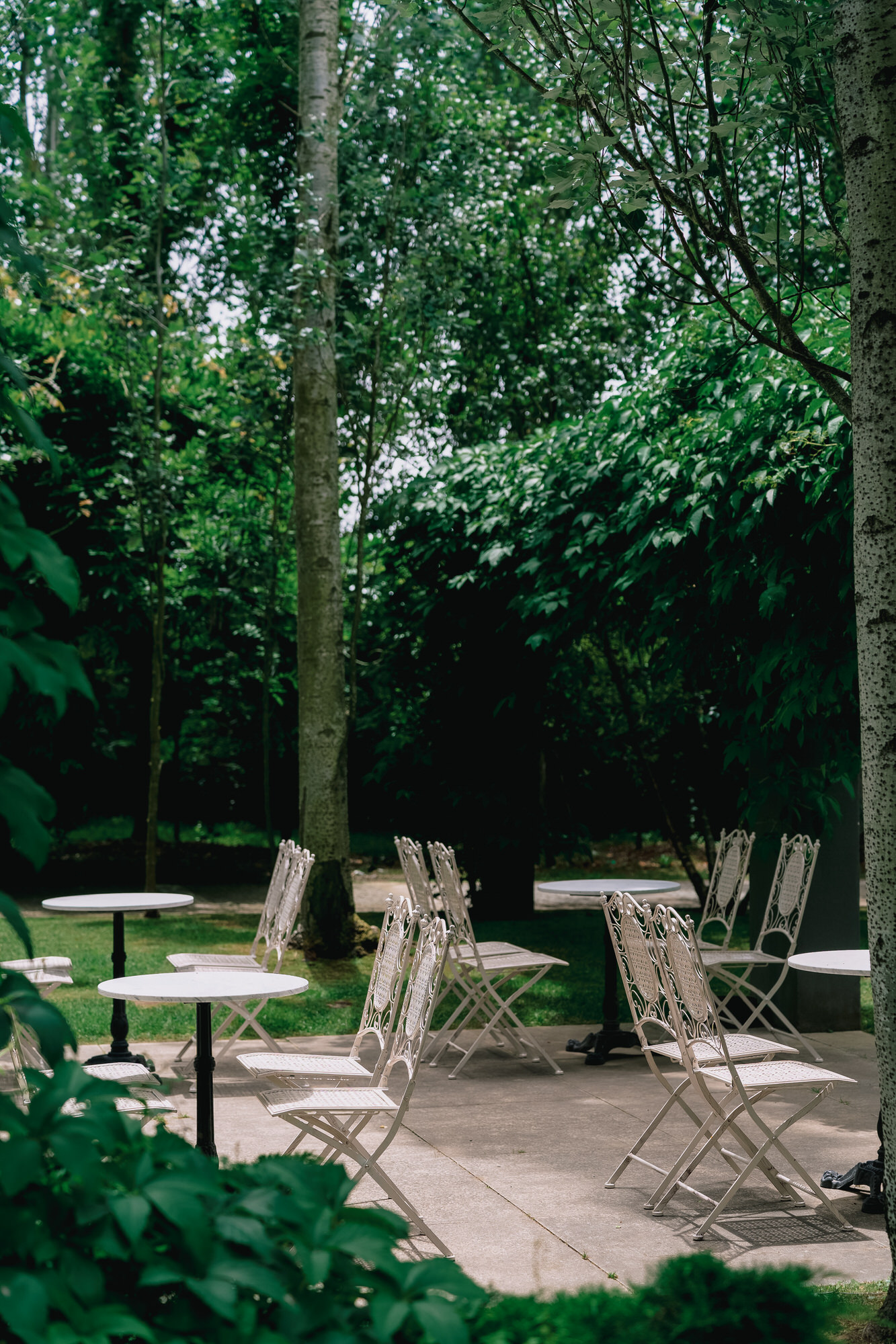 Outdoor garden seating with white metal tables and chairs among trees and greenery, shaded and tranquil