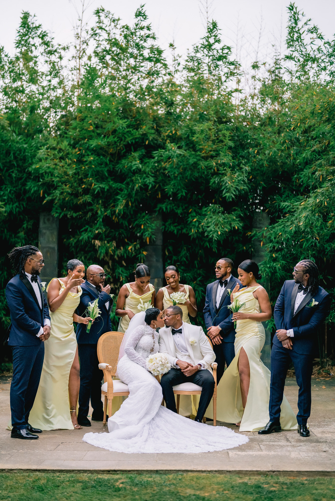 Bride and groom seated center, surrounded by bridesmaids in pale yellow dresses and groomsmen in navy suits at an outdoor wedding, with tall green hedges behind.