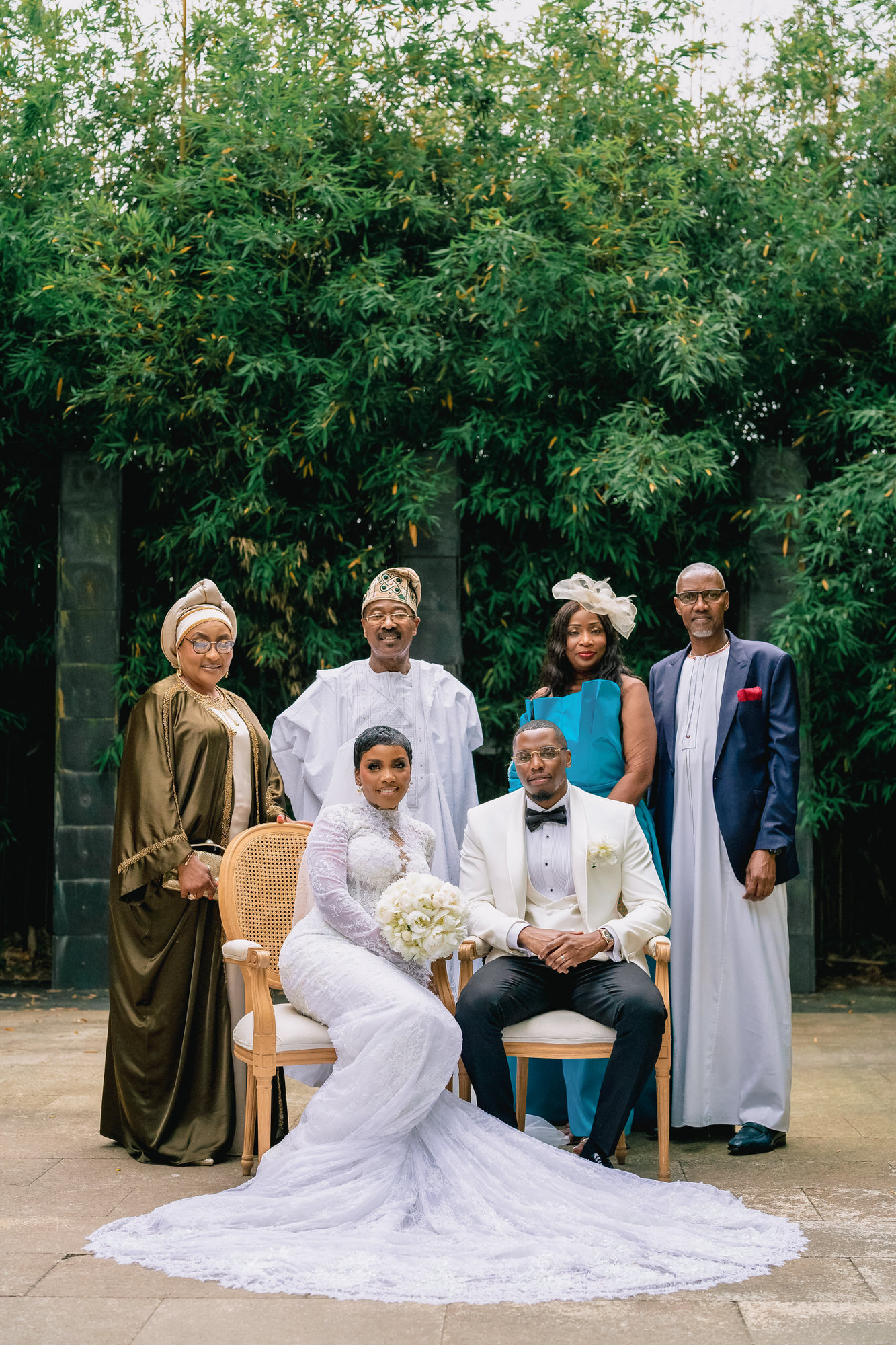 Bride in a white lace gown and veil sits beside a groom in a white jacket and black bow tie, with family behind them outdoors.