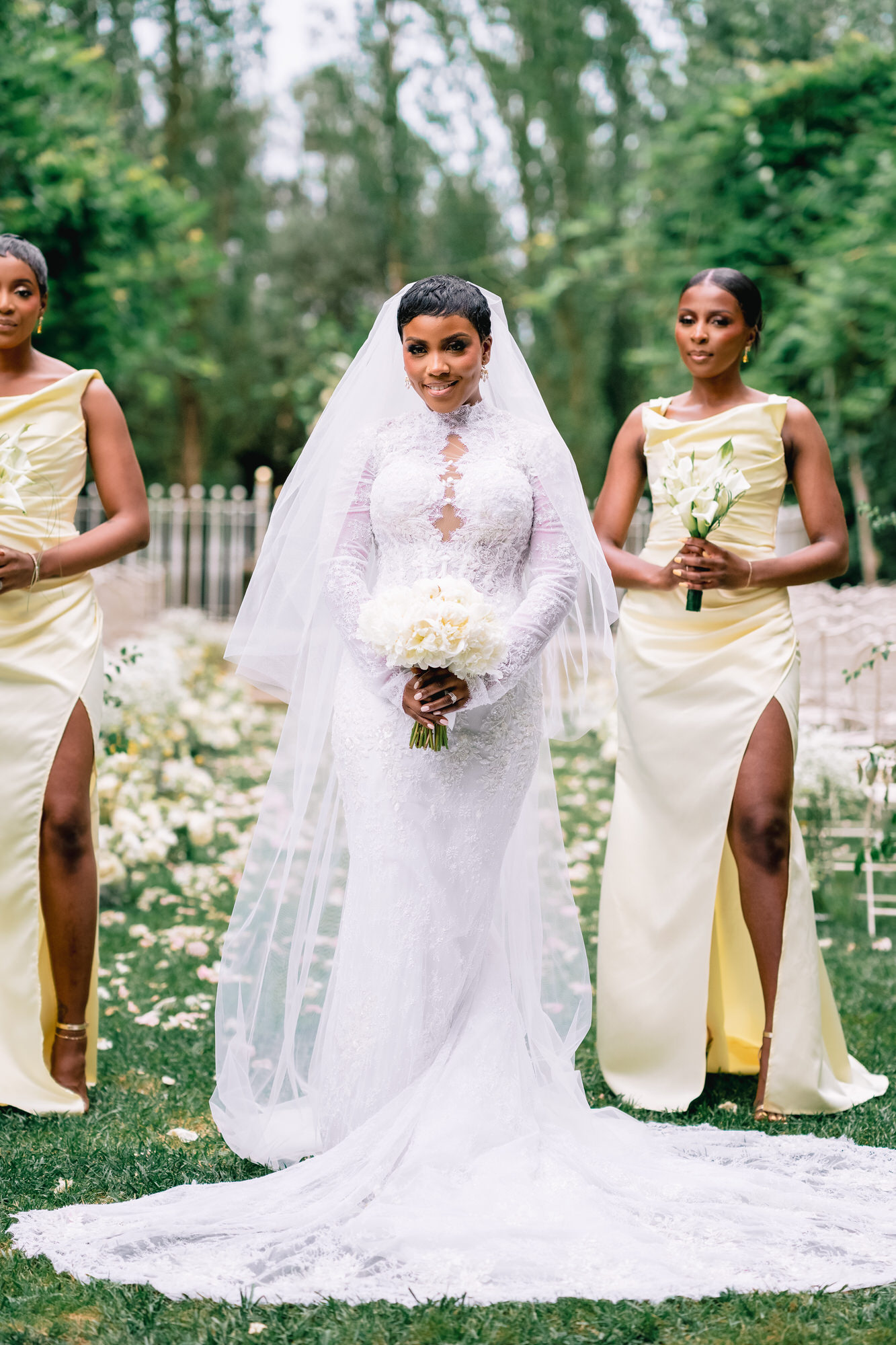 Bride in a white lace wedding gown and veil holding a bouquet, flanked by bridesmaids in pale yellow dresses outdoors.