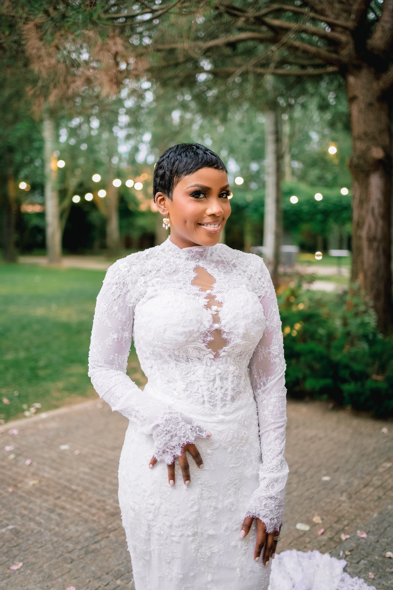 Bride in a white lace wedding gown posing outdoors, smiling with blurred garden lights in the background.