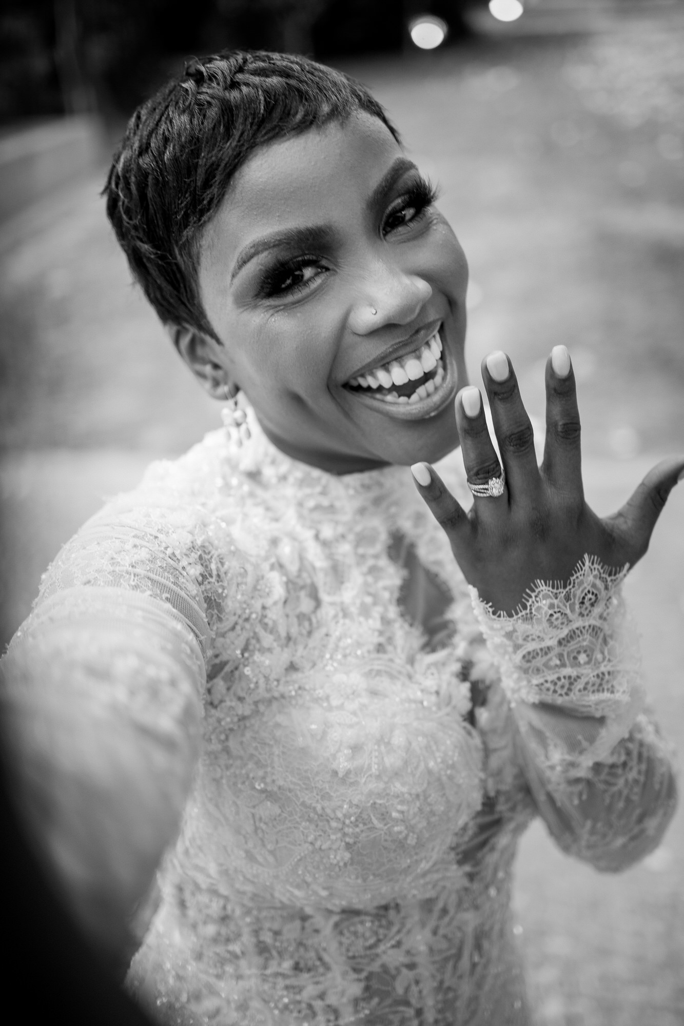 Black-and-white close-up of a smiling bride with short hair, wearing a lace wedding dress and showing rings on her left hand.