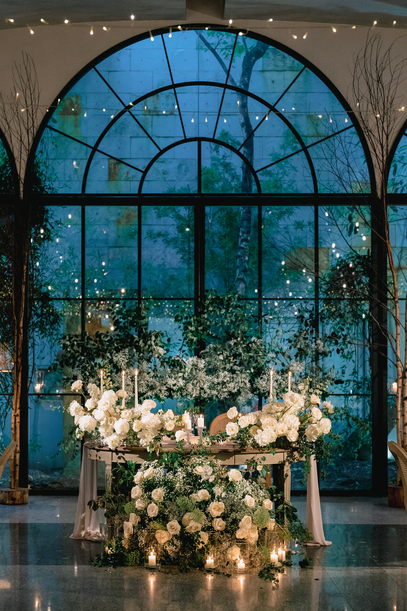 Bride in a lace wedding gown and veil, holding a bouquet of white flowers, next to a groom in a white tuxedo outdoors.