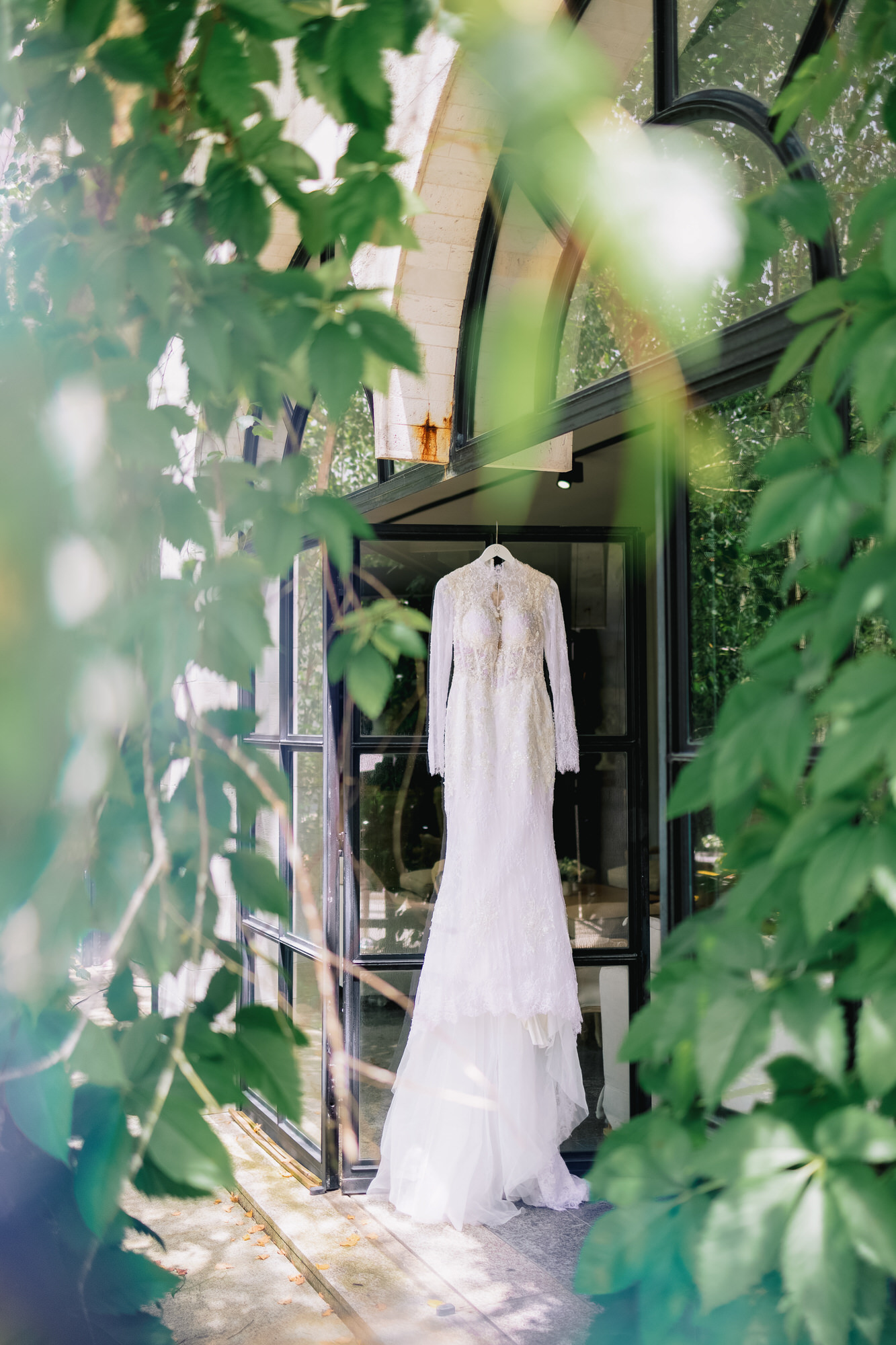 White lace wedding gown hanging in a glass doorway, framed by lush green leaves outside.
