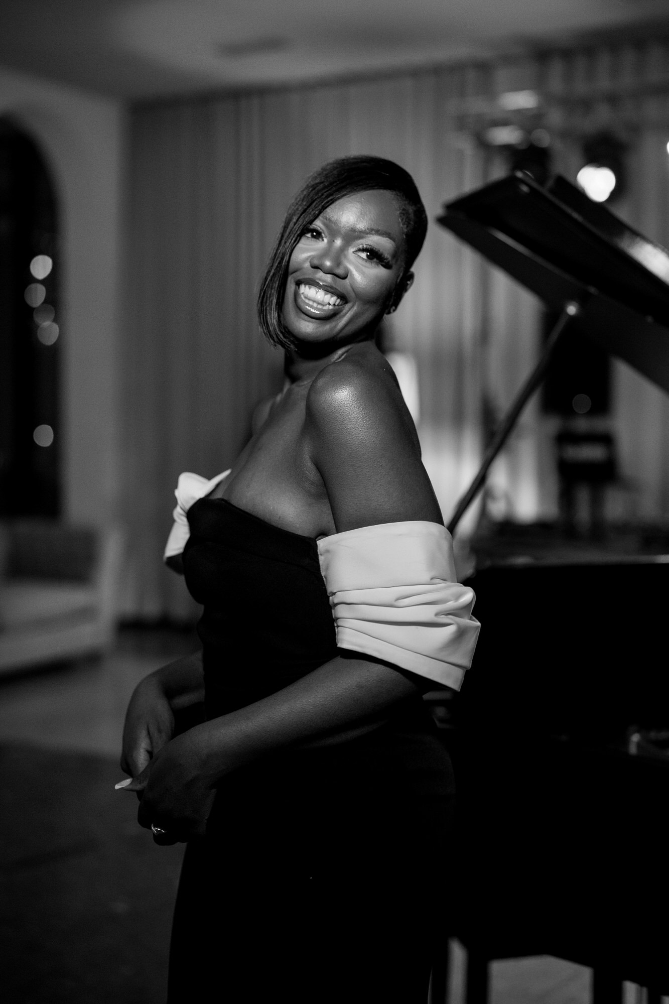 Joyful woman in a black off-shoulder dress with white sleeves posing beside a piano in a dim room, looking at the camera and smiling widely.