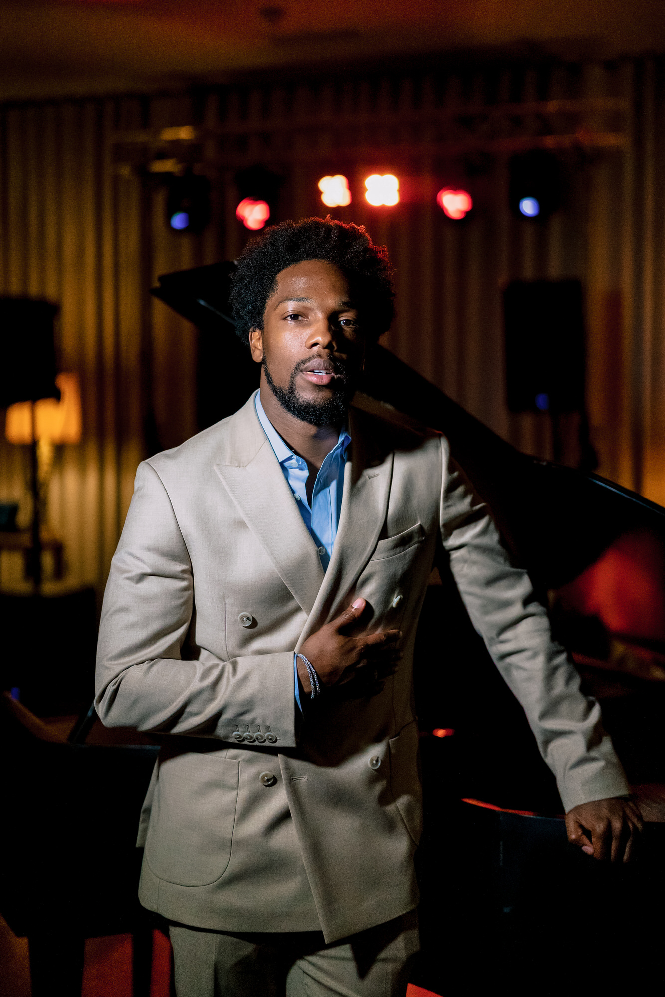 Man in a light beige suit at a piano on a dimly lit stage with colorful stage lights in the background, looking toward the camera.