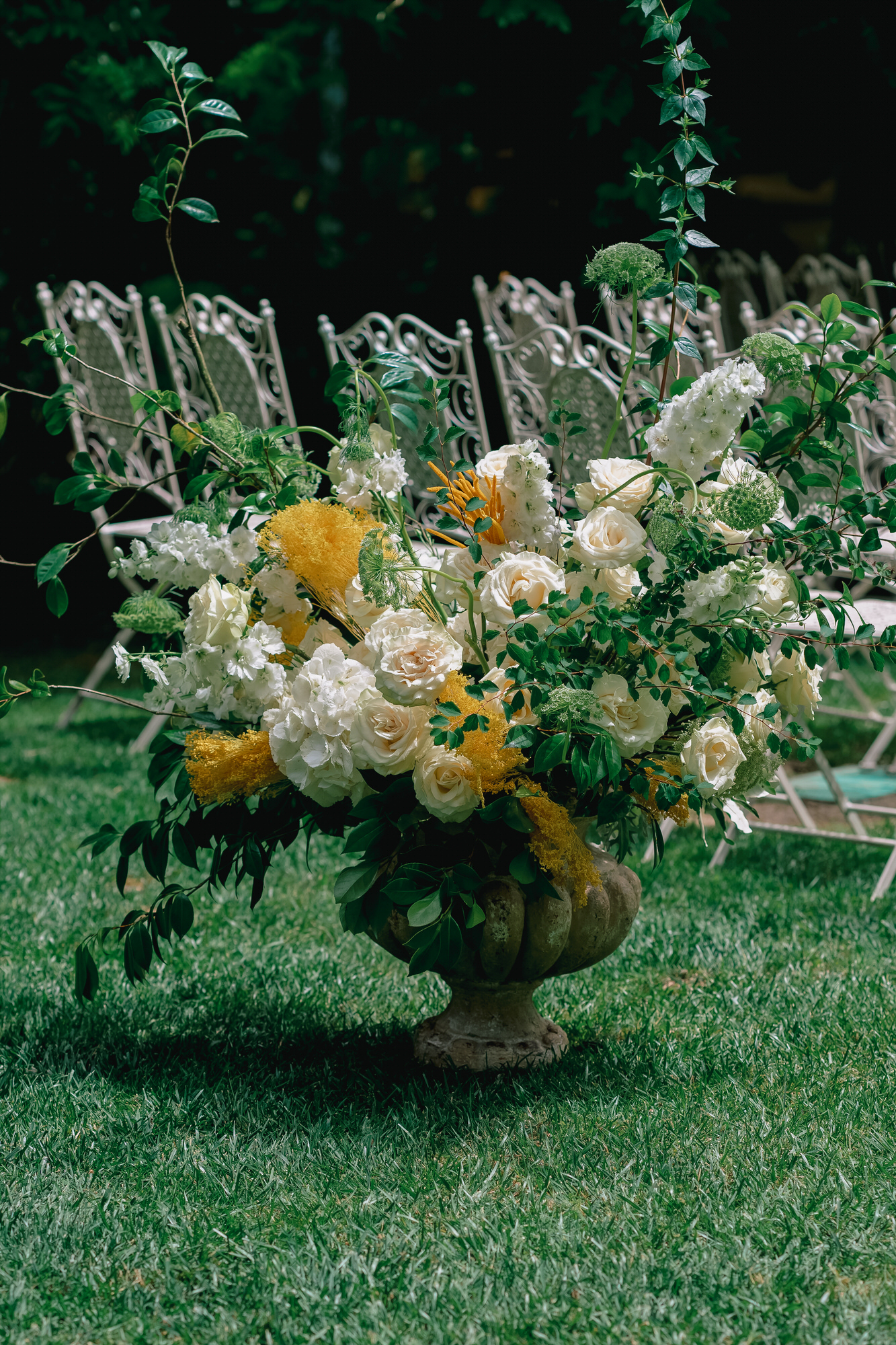 Outdoor wedding centerpiece: white roses and greenery in a stone urn on a grassy lawn, with ornate metal chairs in the background.