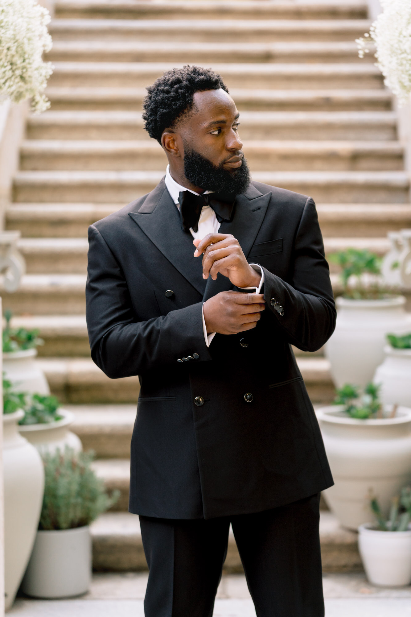 Man in a black tuxedo adjusting his cufflinks on an outdoor stone staircase with potted plants nearby
