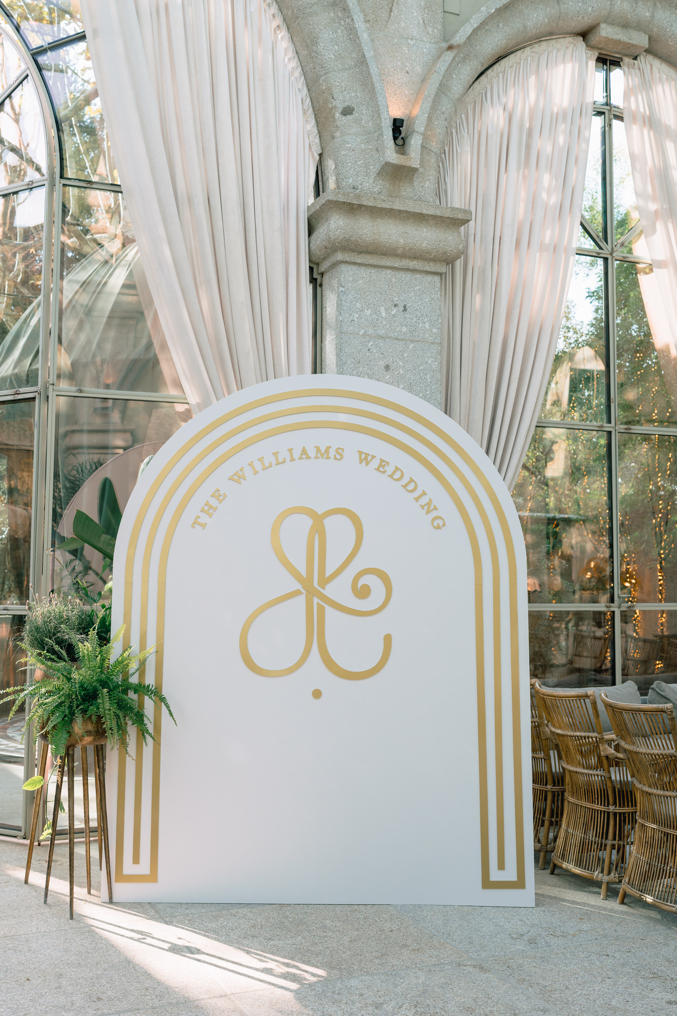 Wedding sign: white panel with gold rings and monogram, 'The Williams Wedding', in front of arched glass windows with white curtains.