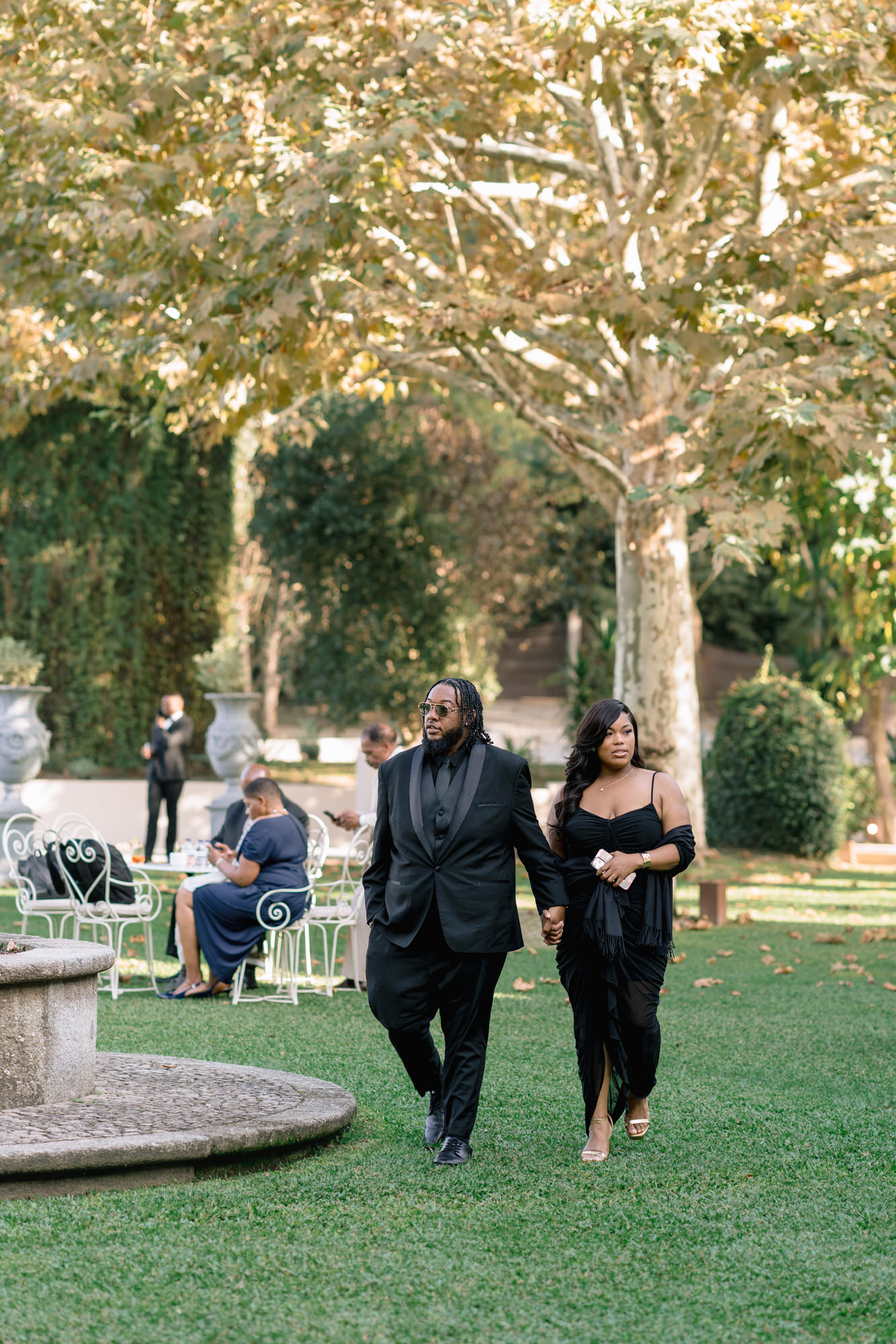 A well-dressed couple walks hand in hand on a grassy lawn at an outdoor event, with guests seated in the background.