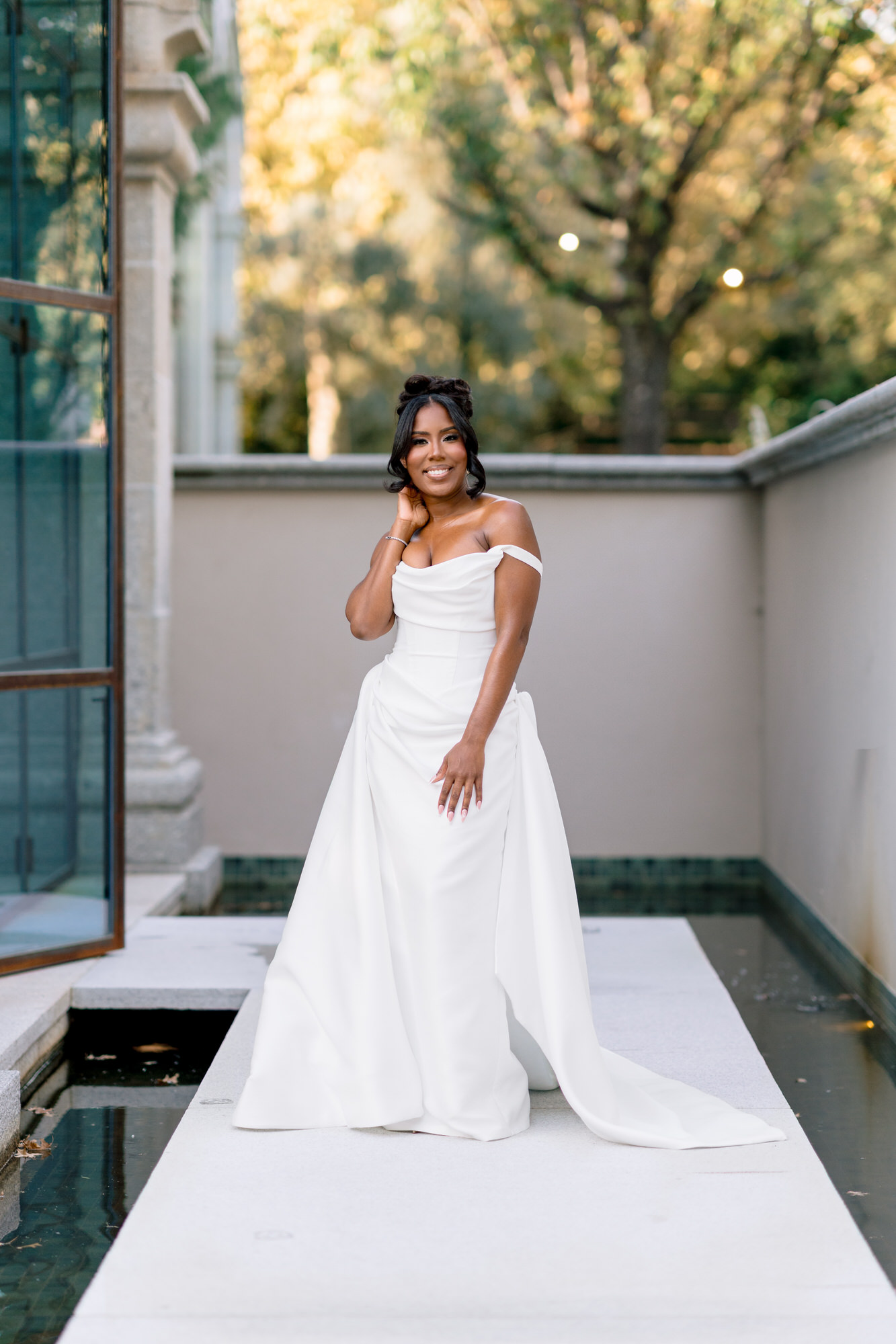 Smiling woman in a white off-the-shoulder wedding dress posing on a light stone walkway beside a shallow reflecting pool outdoors.
