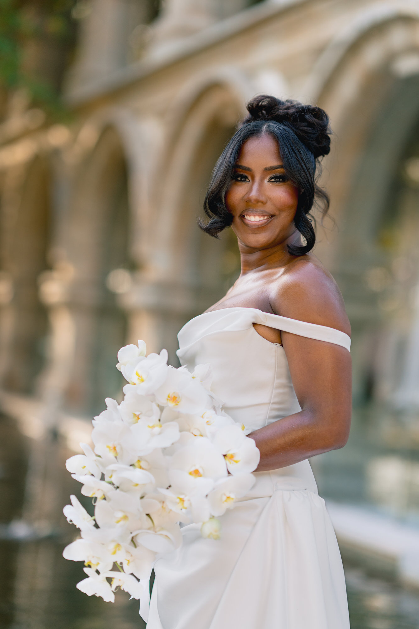 Bride in an off-shoulder white gown smiles, holding a bouquet of white orchids outdoors.