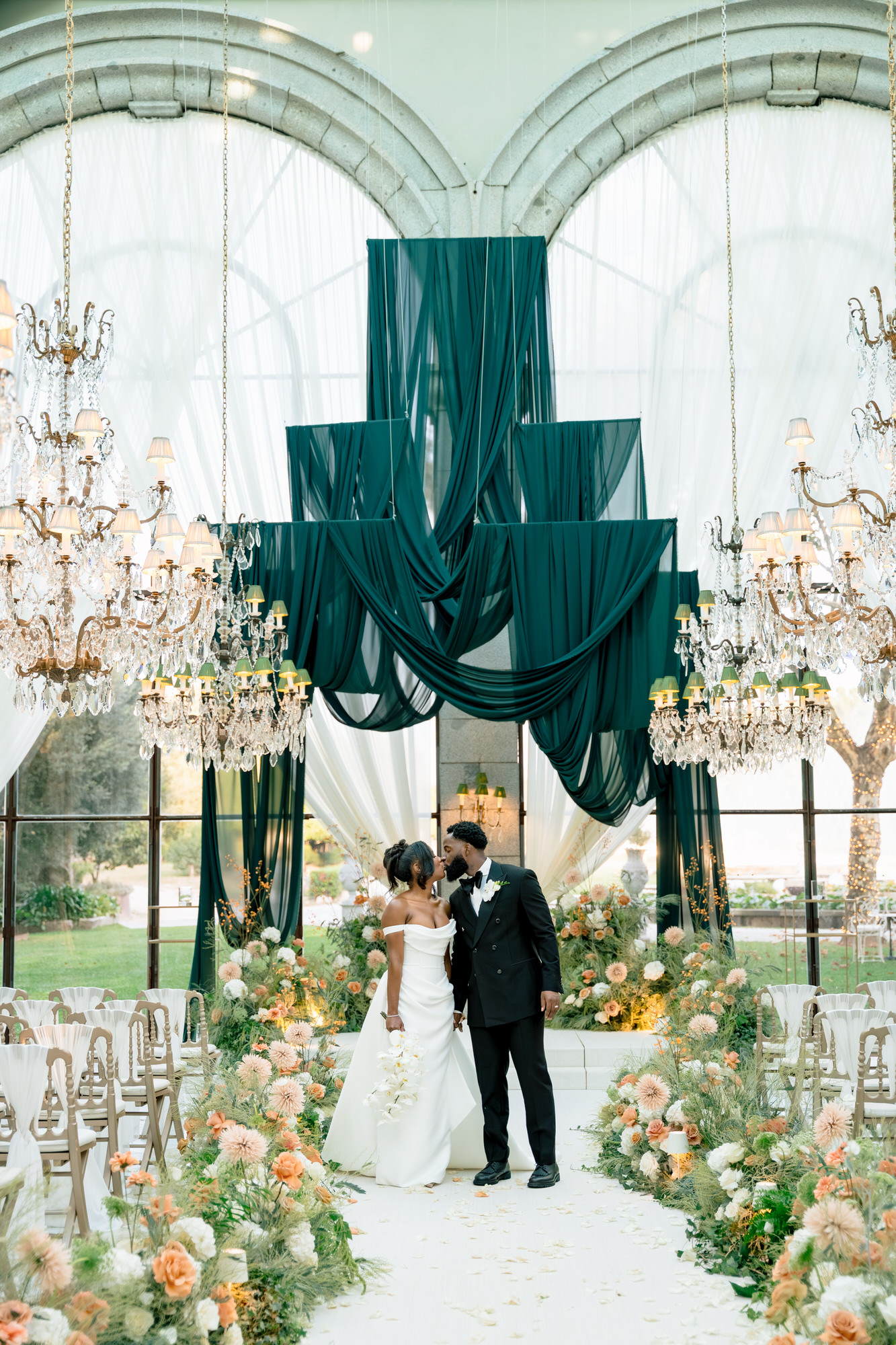 Bride and groom share a kiss beneath teal draped arches in a grand, chandelier-lit wedding venue.