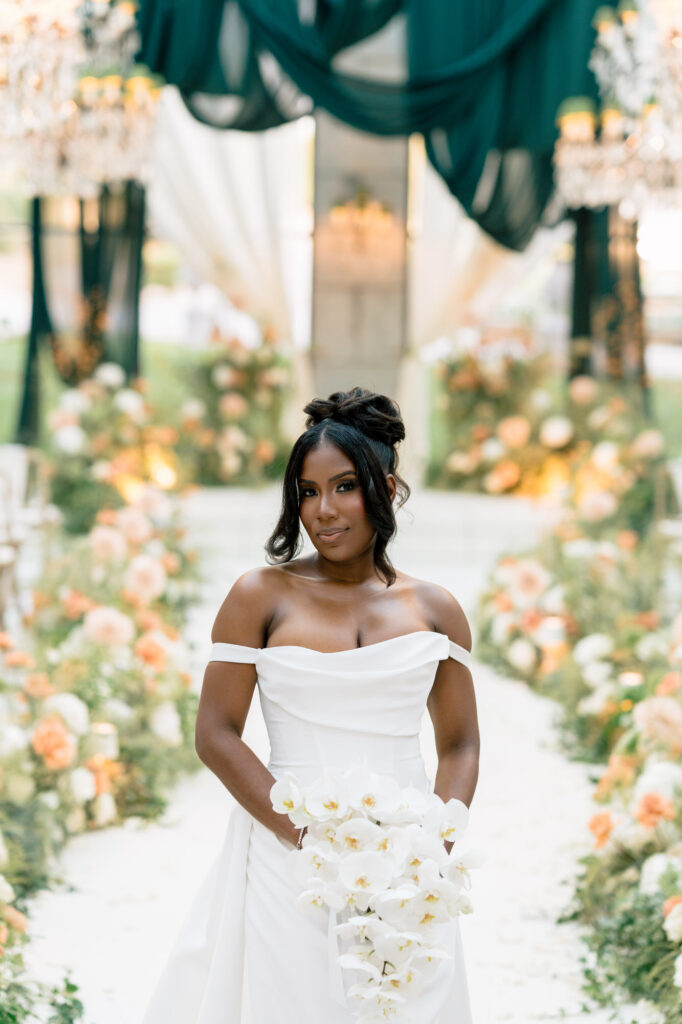 Bride in off-shoulder white gown holding a cascading bouquet of white orchids at a floral-filled outdoor wedding aisle.