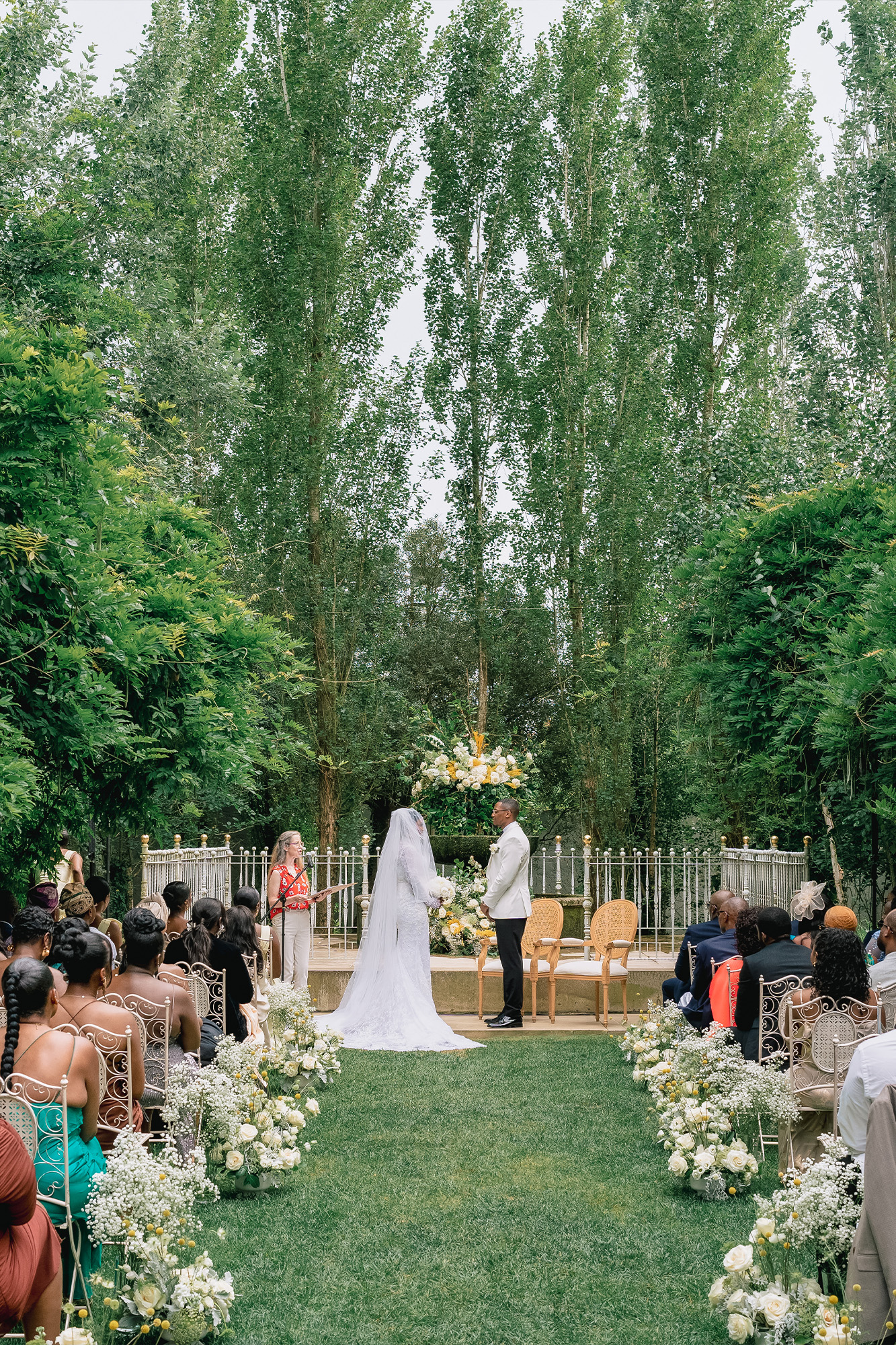 Outdoor wedding ceremony with the bride and groom facing each other amid lush greenery, guests seated along a flower-lined aisle.