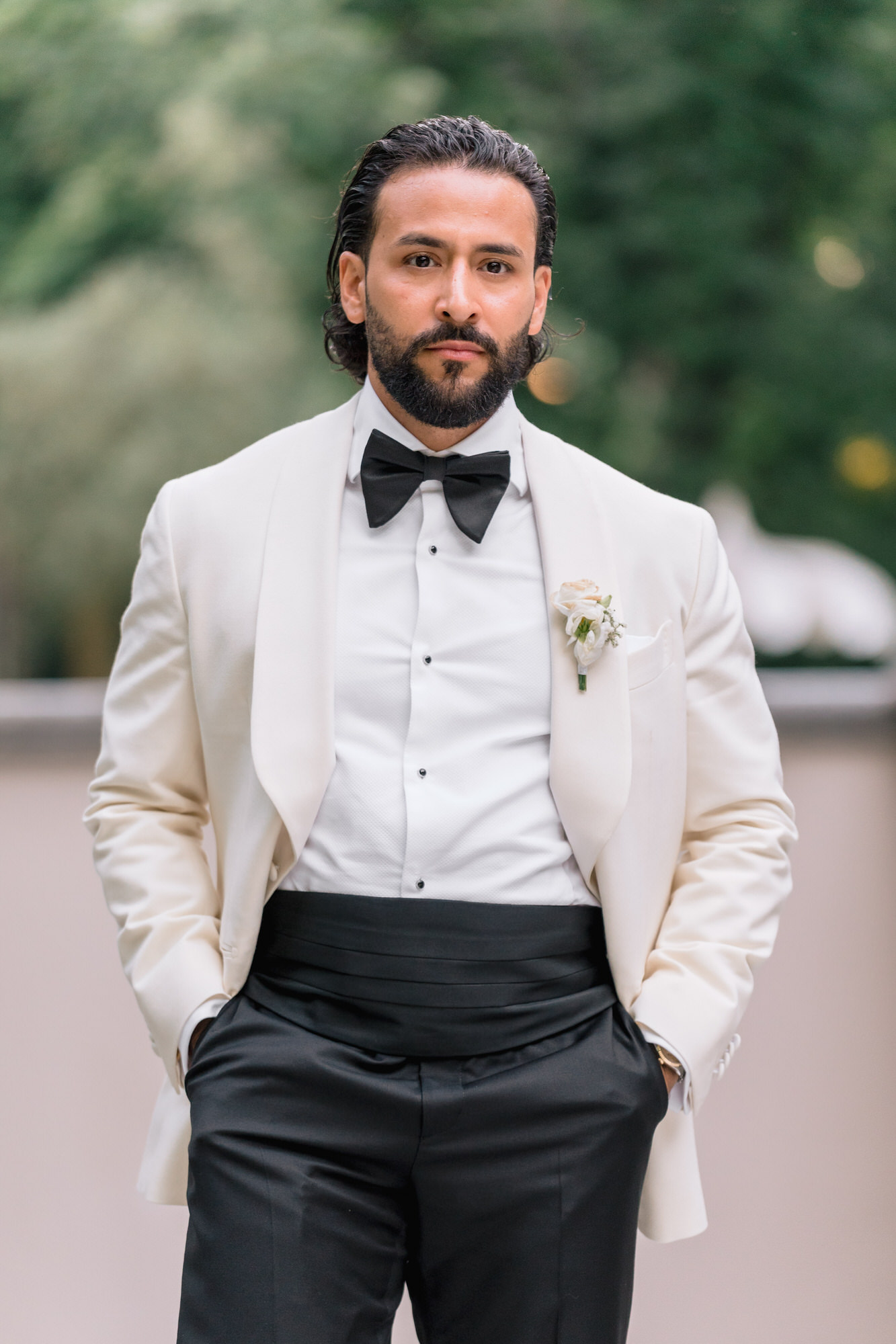 Portrait of the groom in a cream shawl-collar dinner jacket and black bow tie, hands in his pockets, garden behind