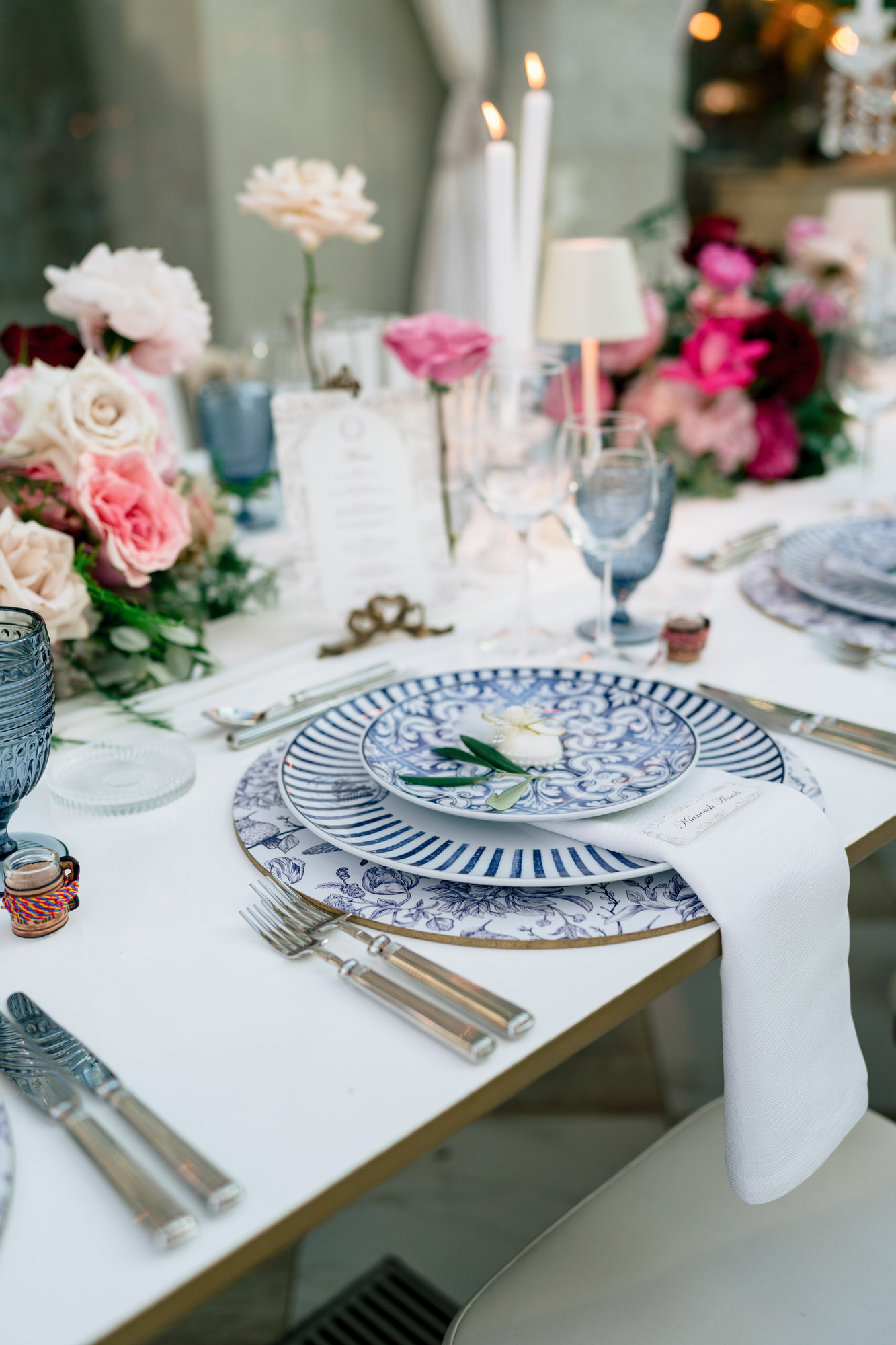 A place setting with a blue patterned plate, white napkin and pink roses, candles behind