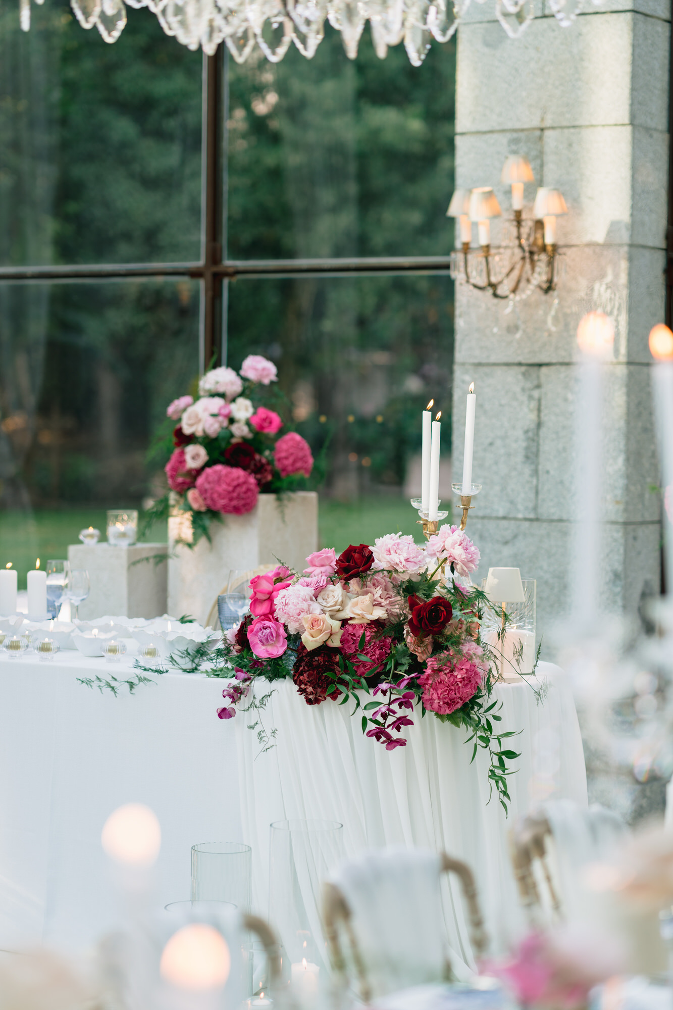 Pink and burgundy floral arrangement on a white table beside tall windows, candles lit