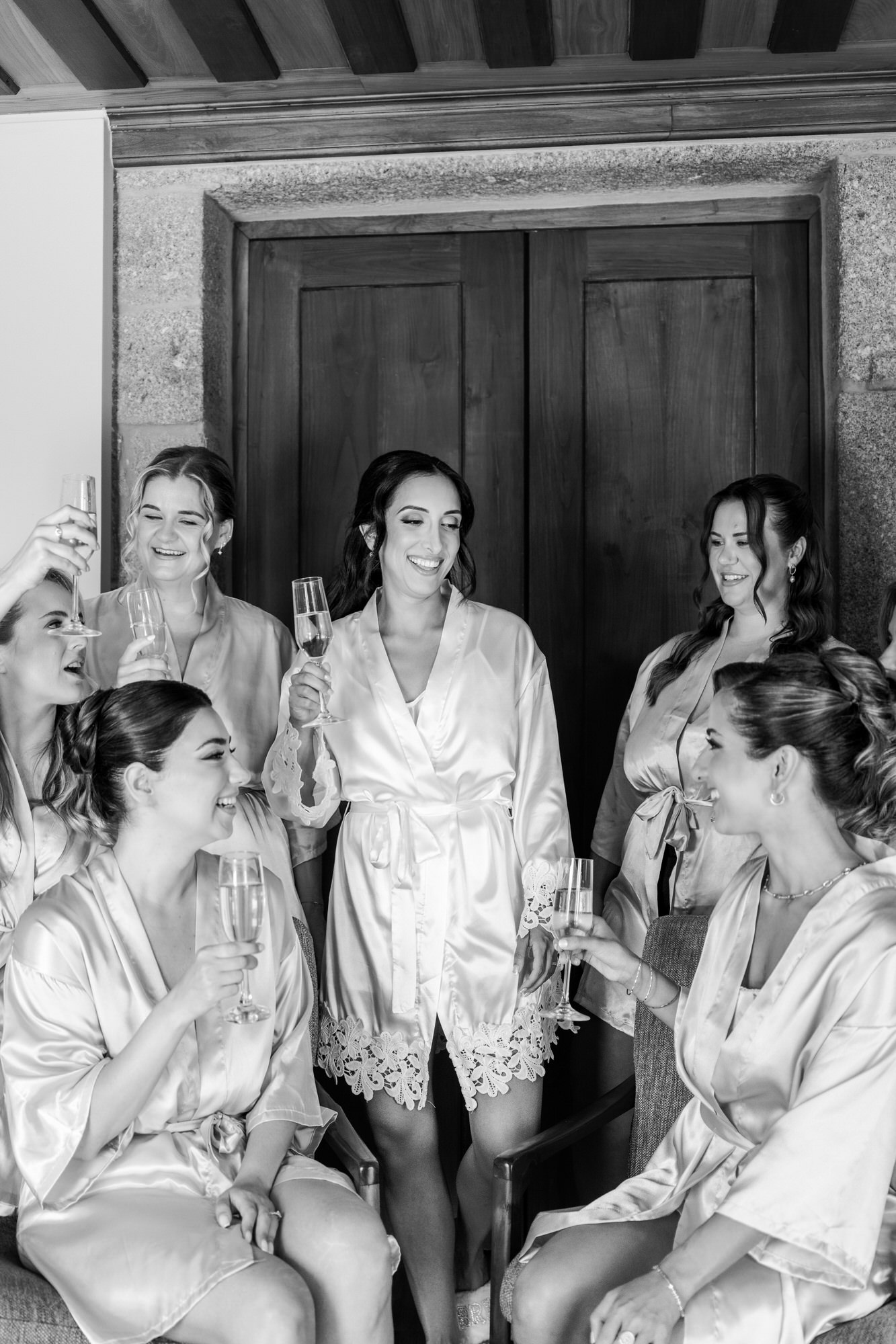 Black-and-white portrait of the bride and her bridesmaids in matching robes, raising glasses in front of tall wooden doors