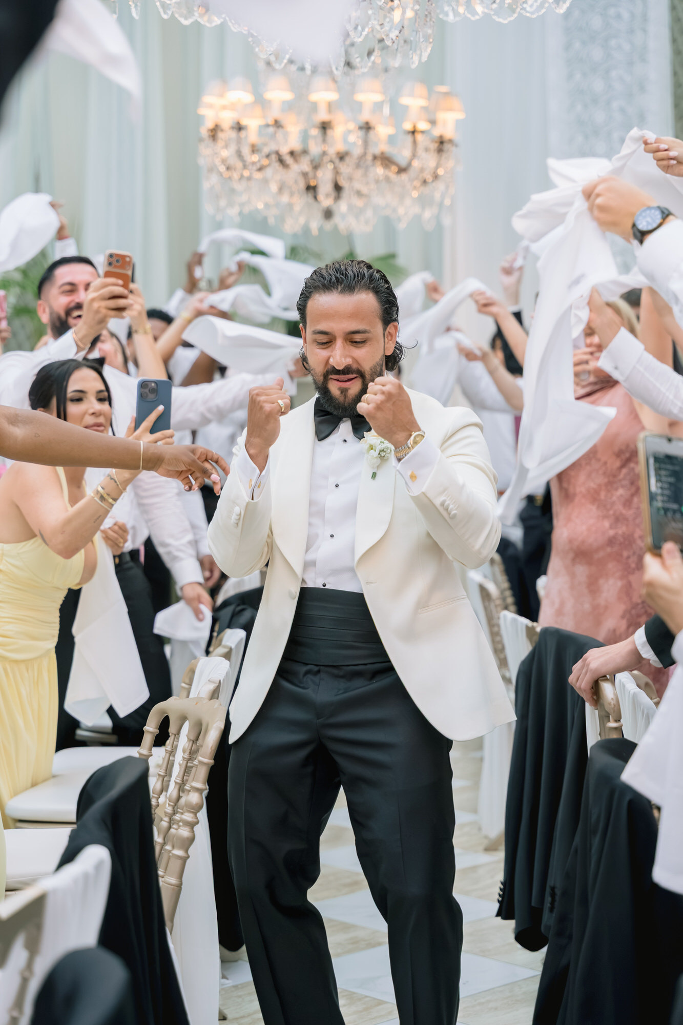 The groom dancing among guests who wave white napkins beneath a crystal chandelier