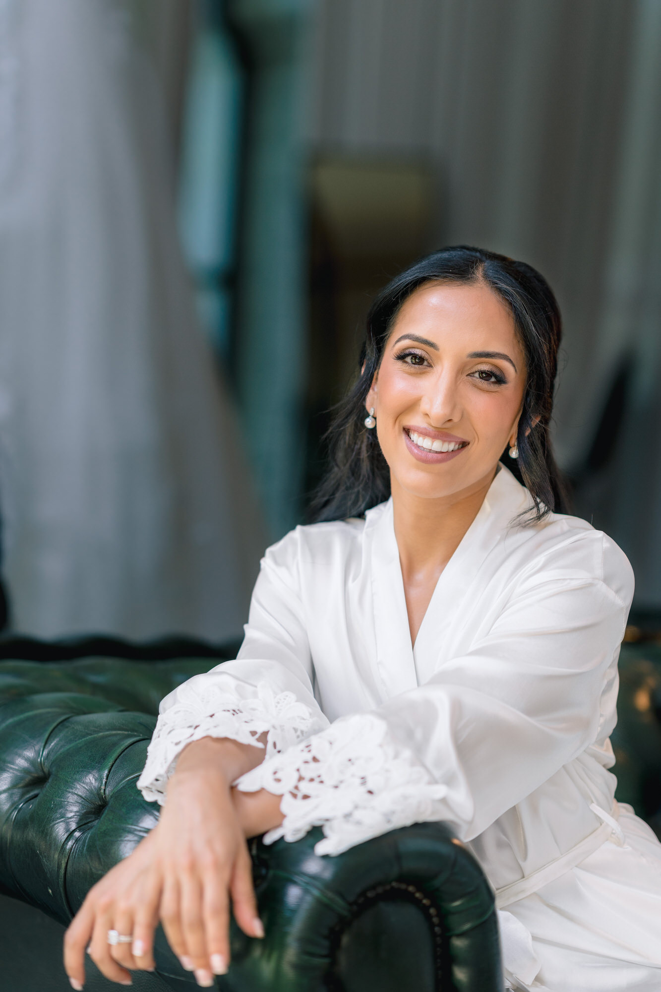 The bride in a white lace robe, smiling as she rests her arm on a green leather armchair