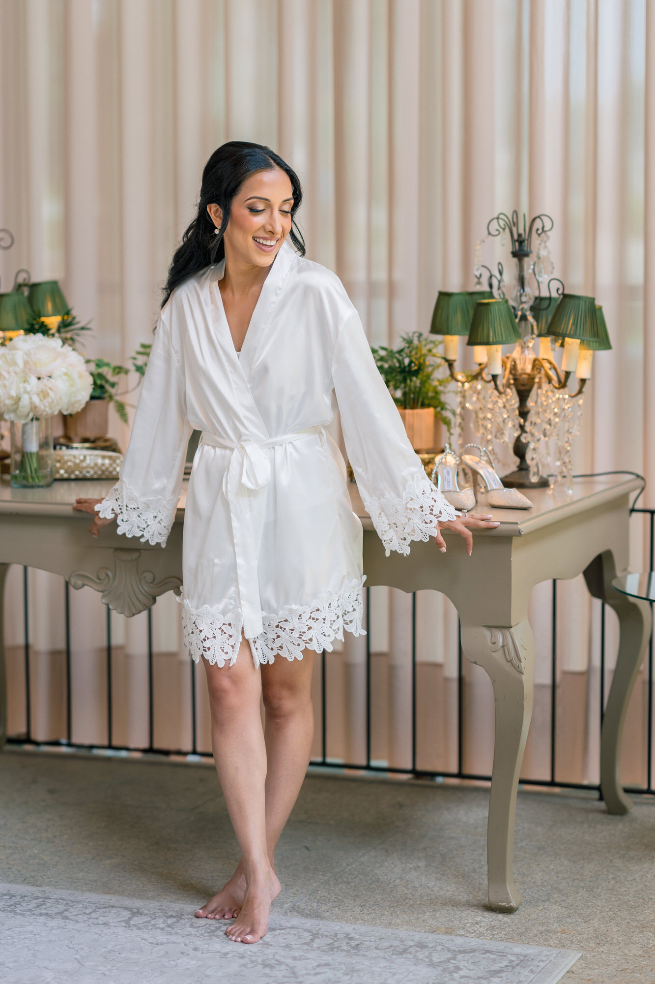 The bride standing in a white lace robe beside a console table with a lamp and fresh flowers