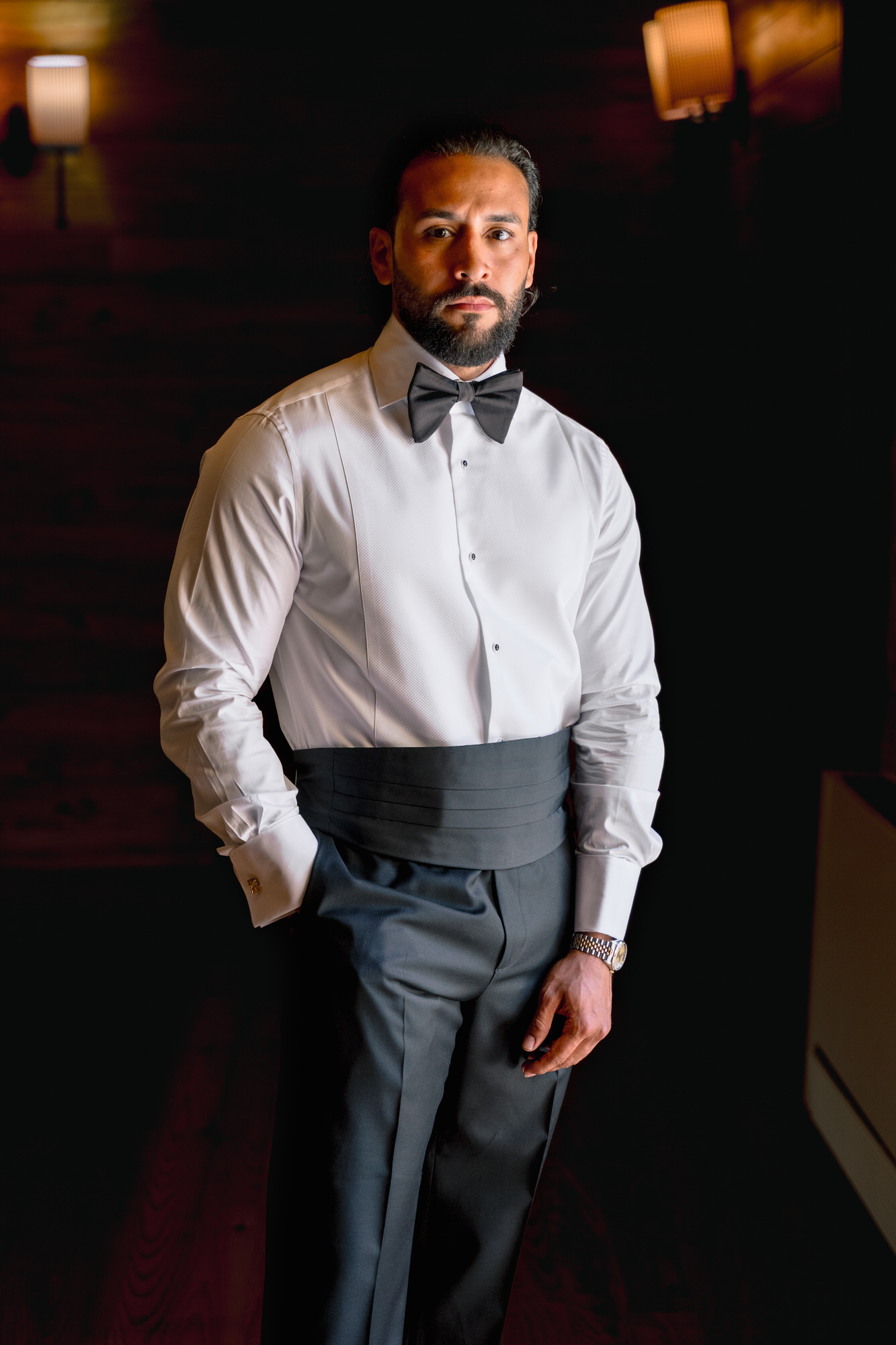 The groom in a white shirt, bow tie and grey cummerbund, standing in a dimly lit room before the ceremony
