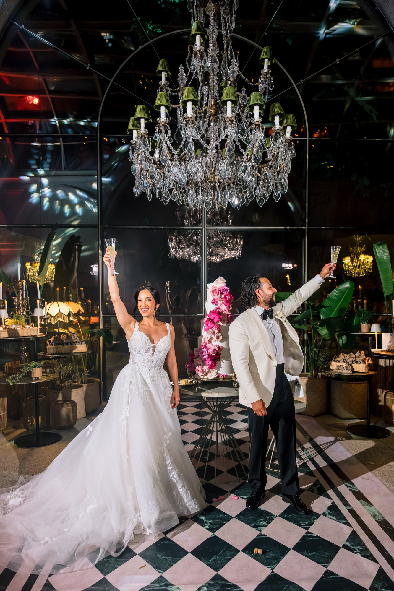 Hana and Andres celebrating with their arms raised on a black-and-white checkered floor beneath a chandelier