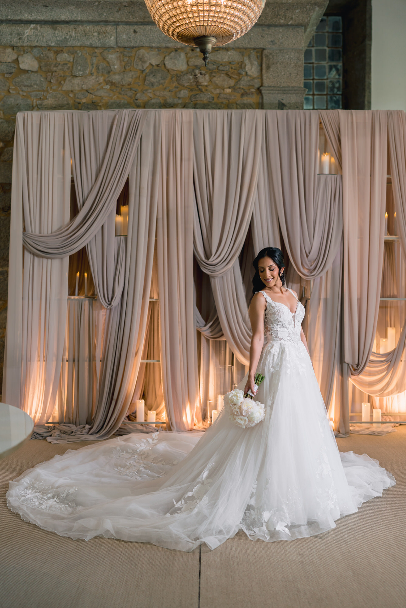 Full-length portrait of the bride, her long train spread out before draped curtains and candles
